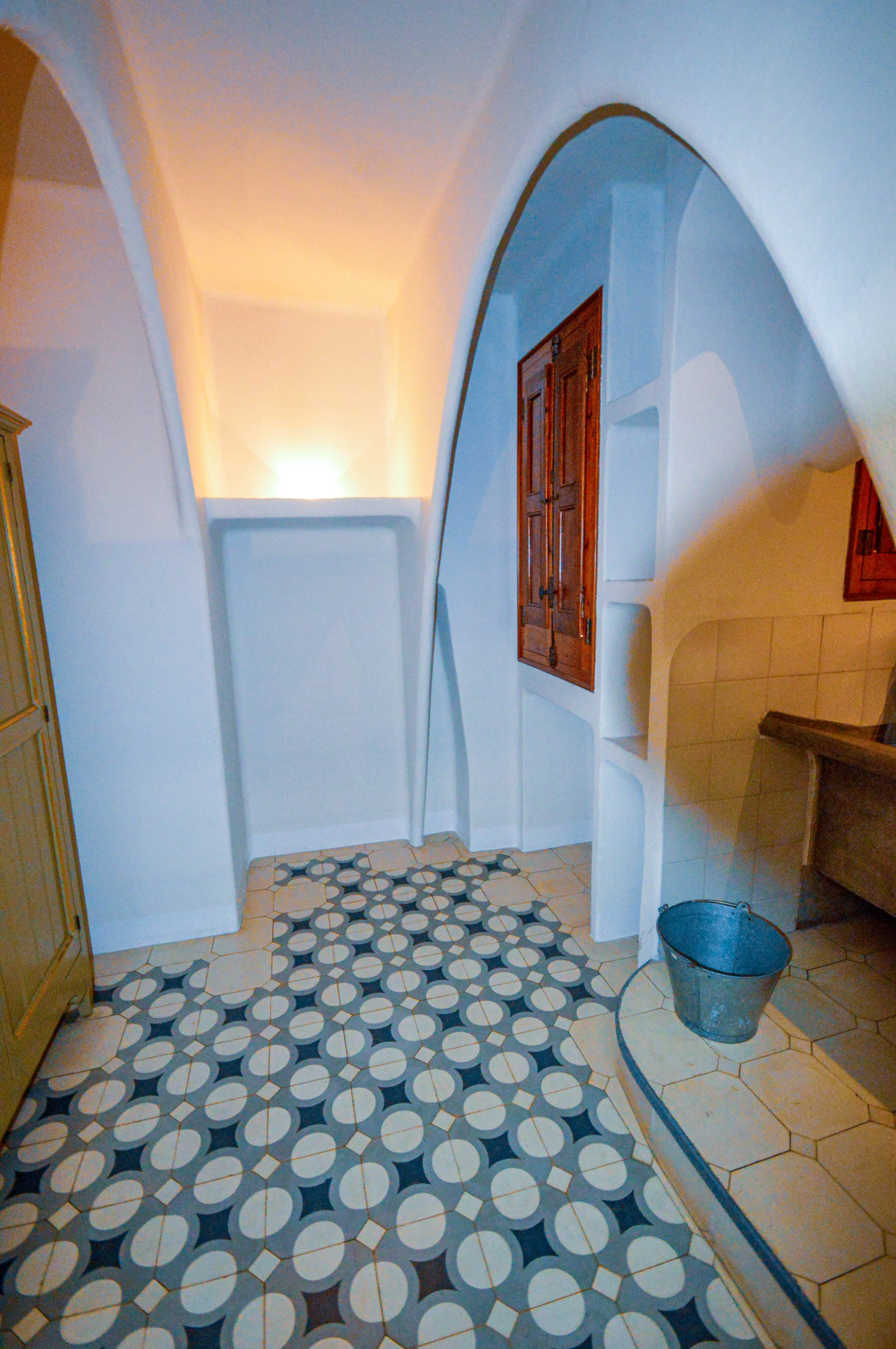 The image depicts an interior space with a unique architectural design. The floor features a patterned tile design with circles and lines, while the walls are painted white. There is a wooden door with metal fittings, and the ceiling has an arched shape. A metal bucket is Barcelonad on the floor near the door. The overall atmosphere suggests a traditional or historic setting.