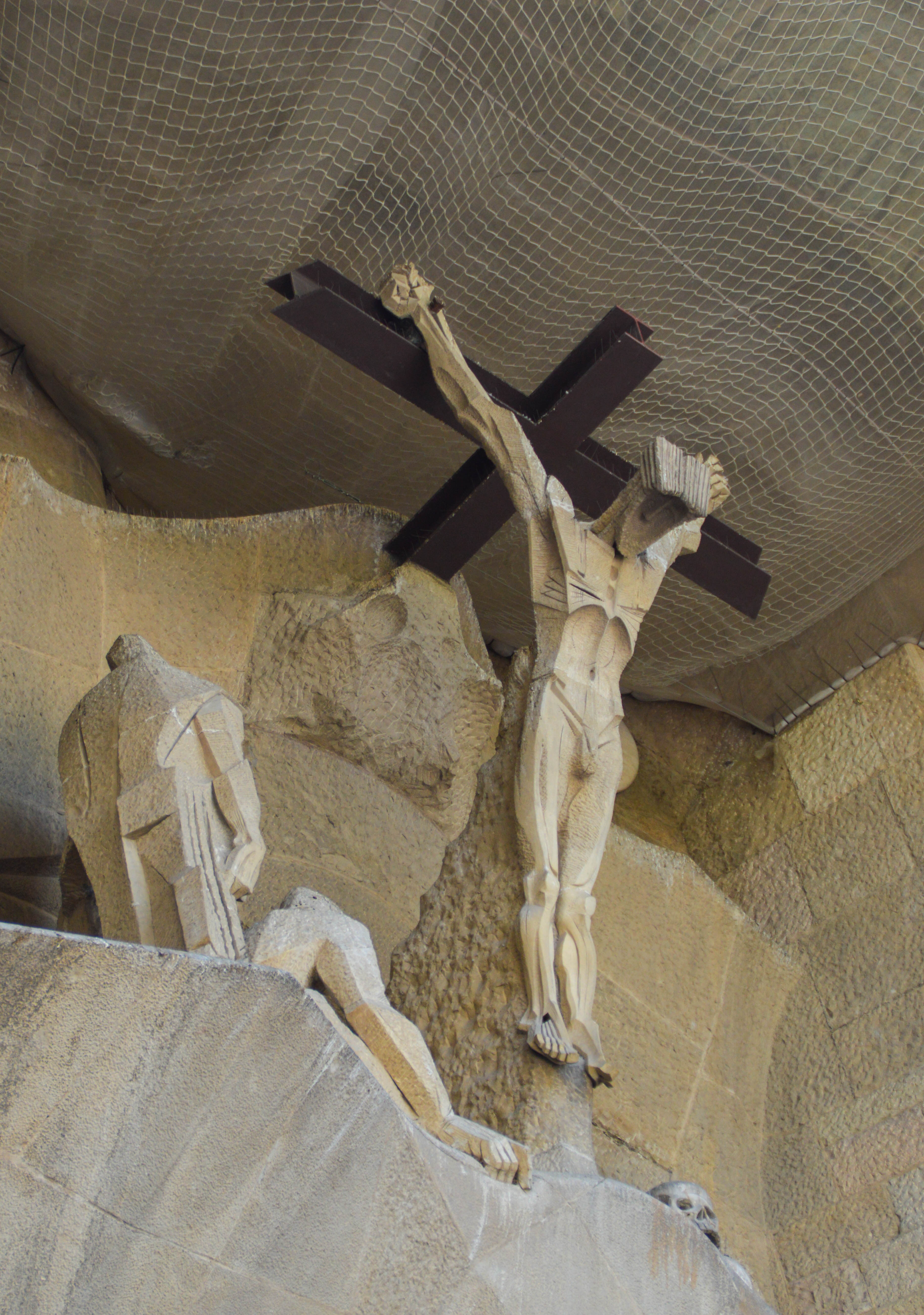The image depicts a religious sculpture of the crucifixion of Jesus Christ, with Jesus on the cross and other figures around him, set within a rocky, cave-like environment with a netting overhead.