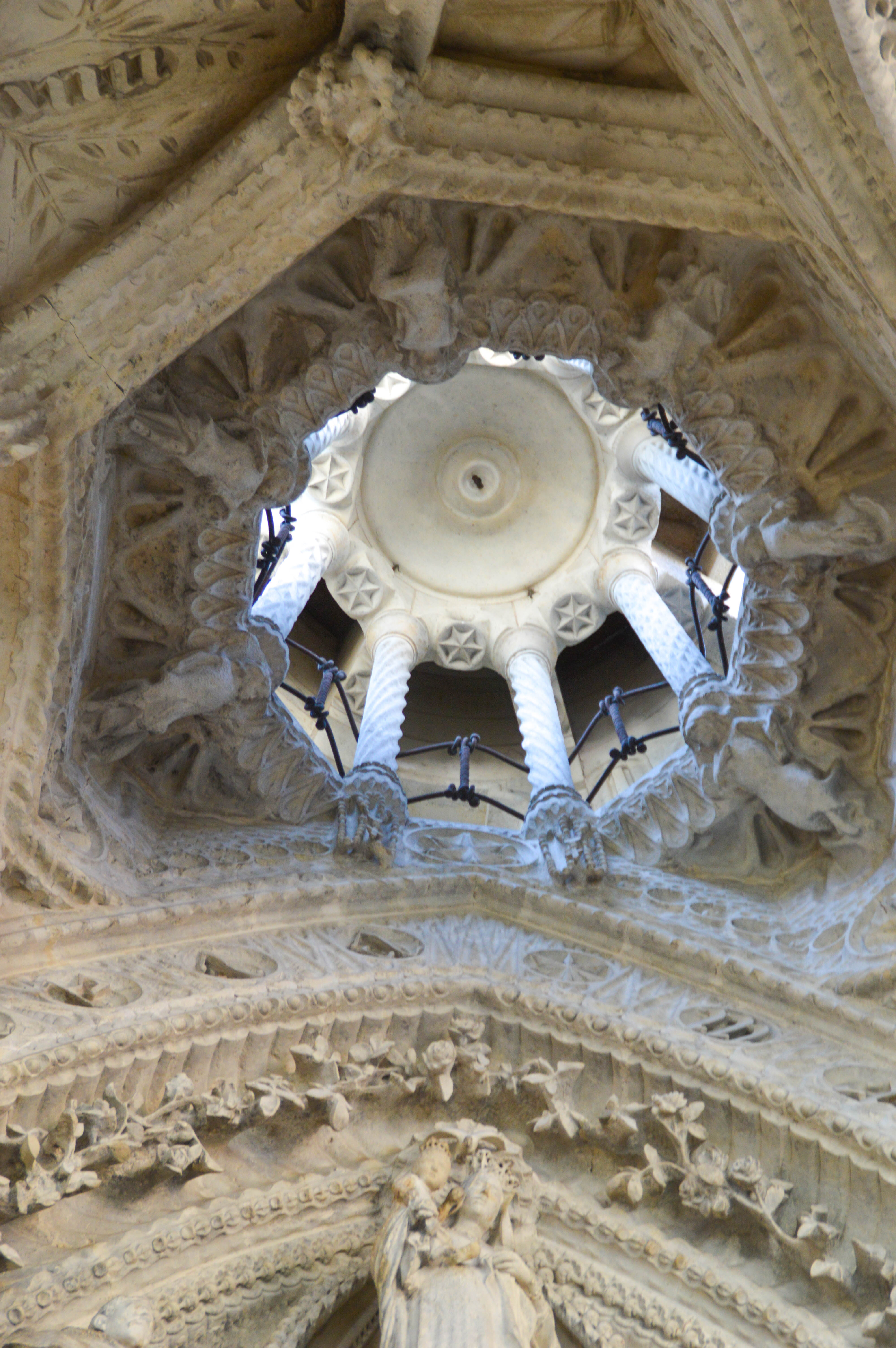 The image depicts an intricately carved stone ceiling, featuring a central circular motif surrounded by detailed floral and geometric patterns. The ceiling includes elaborate sculptures of angels and other figures, with chains hanging from the structure, adding to the ornate design.