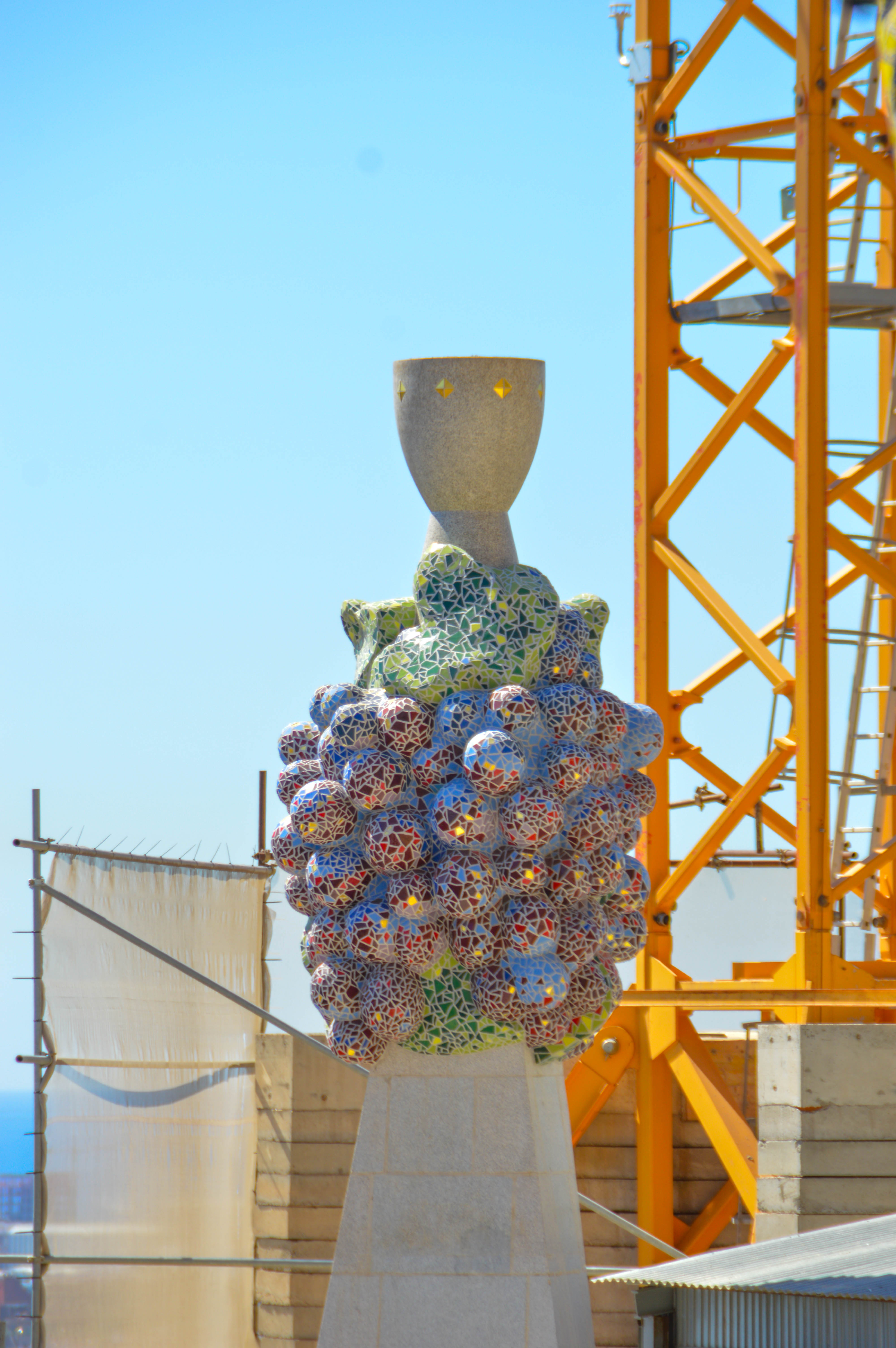 The image shows a colorful mosaic sculpture with a large, ornate base and a simple, wide-mouthed vase on top. The sculpture is situated on a stone pedestal and is surrounded by a construction site with scaffolding and a yellow crane.