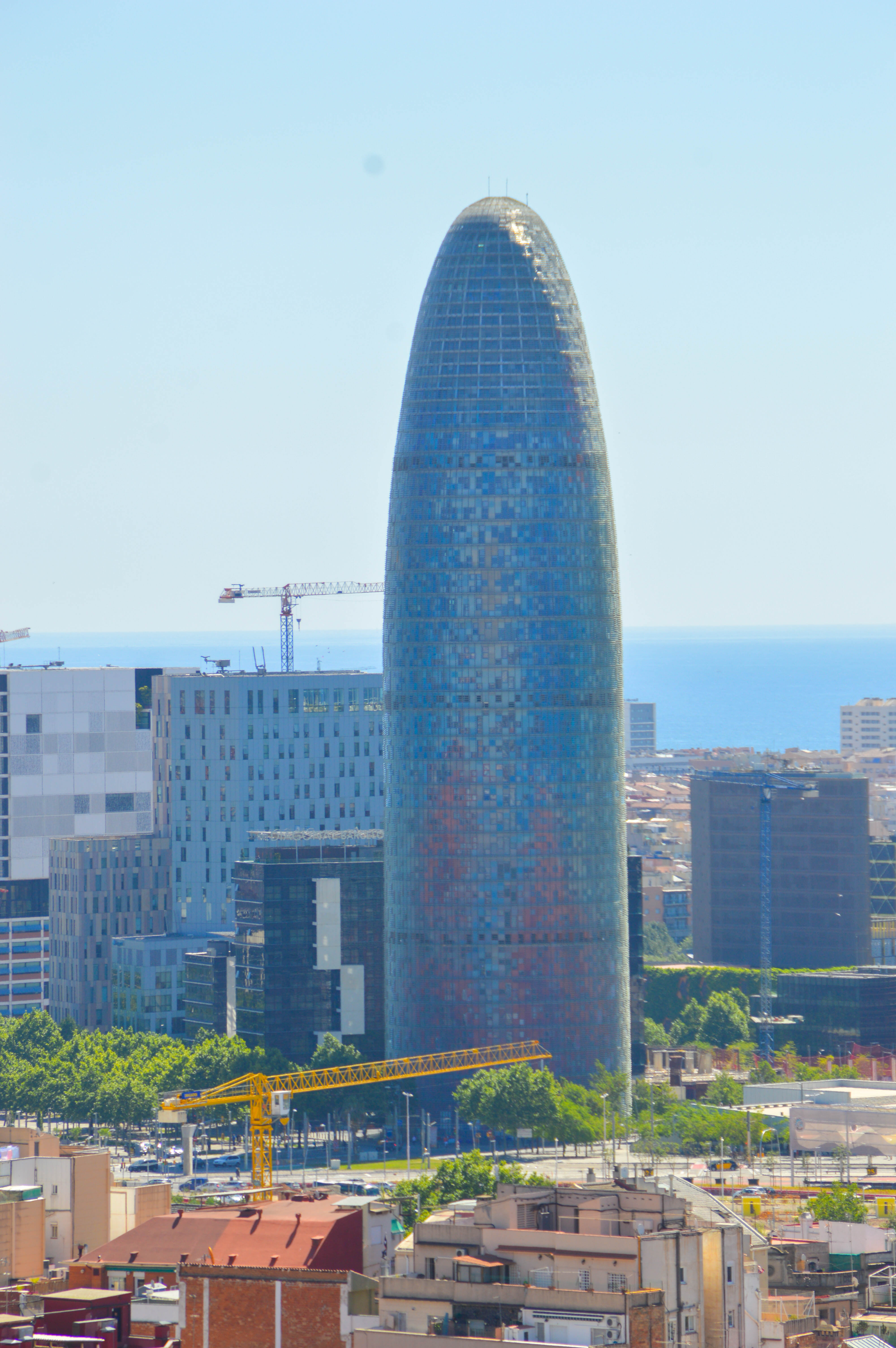 The image depicts a cityscape with a prominent, uniquely designed tall building that resembles a bottle gourd or a vase. This building is surrounded by other modern structures and is situated near the coast, as the sea is visible in the background. There are construction cranes in the vicinity, indicating ongoing development. The area around the tall building includes a mix of older, smaller buildings and green spaces.