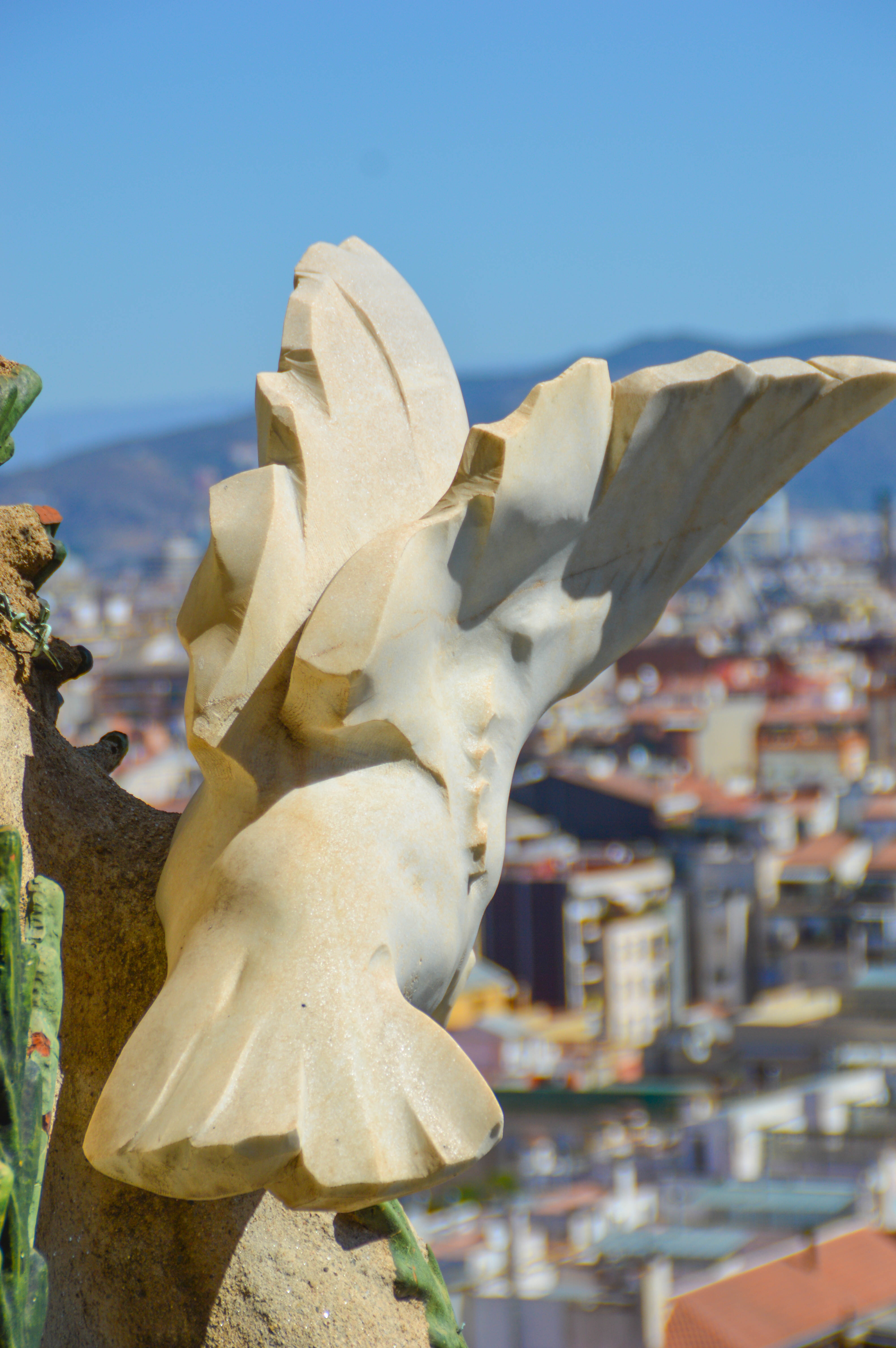 The image depicts a close-up view of a sculpted stone figure, likely a gargoyle, with a cityscape in the background. The figure has a stylized, abstract appearance with angular features and appears to be part of a larger architectural structure. The cityscape in the background shows a mix of buildings with various colors and architectural styles, under a clear blue sky.