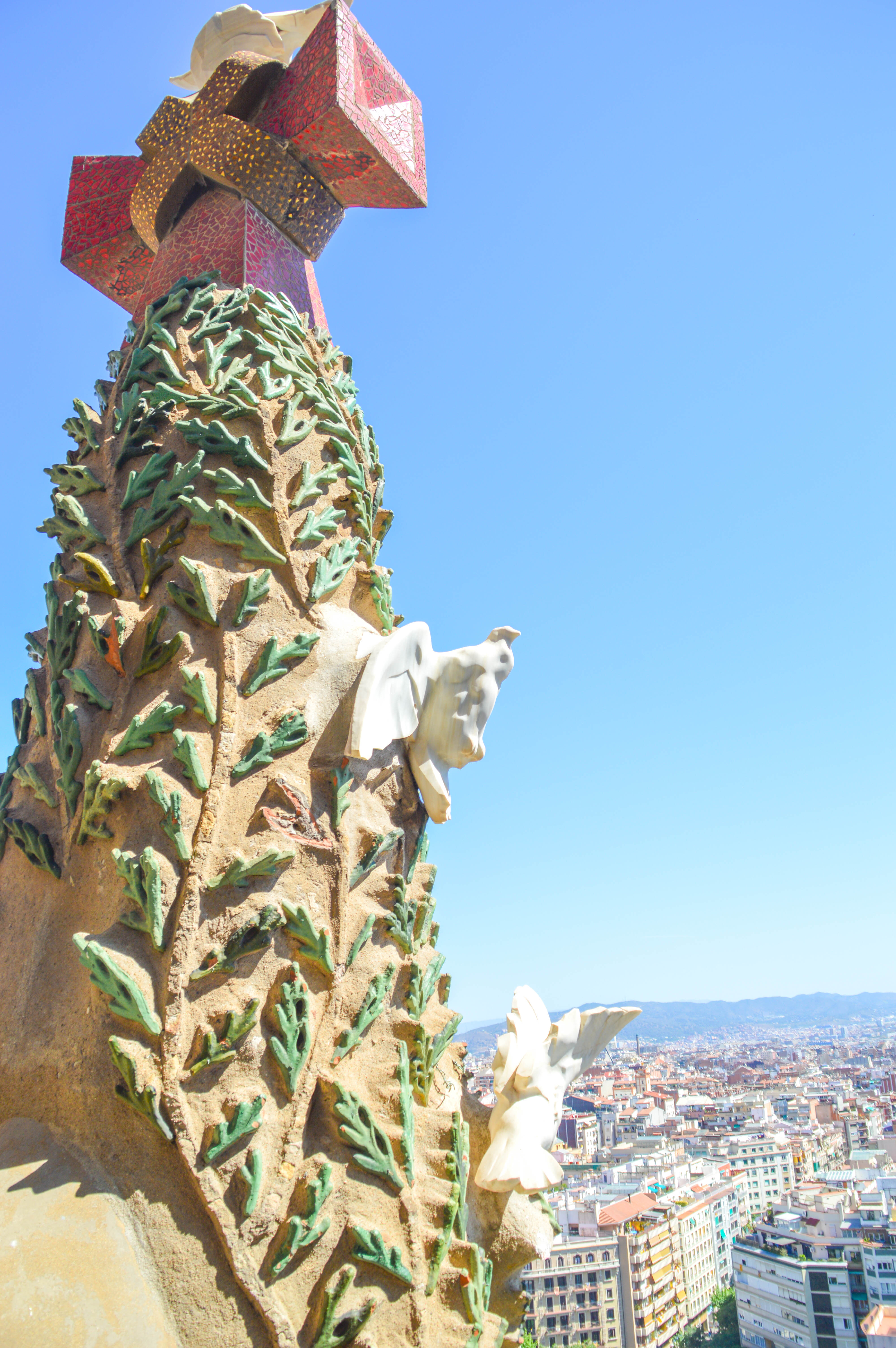 The image depicts a detailed view of the chimney-like decorations on the roof of the Sagrada Familia, a renowned architectural work by Antoni Gaudí located in Barcelona, Spain. The chimneys are adorned with intricate mosaics and sculptural elements, showcasing Gaudí's distinctive and imaginative style. The background reveals a panoramic view of the city of Barcelona under a clear blue sky.