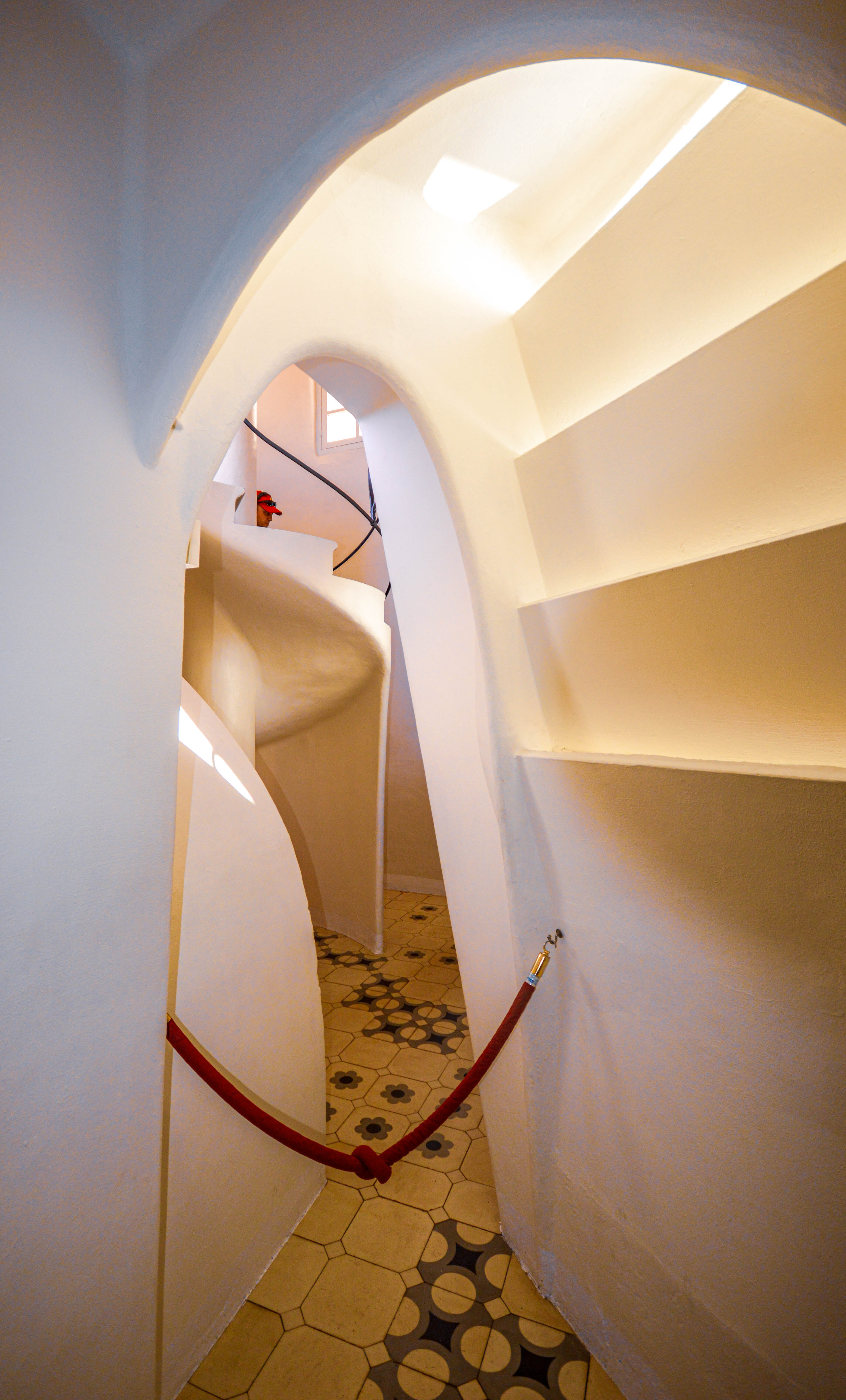 The image depicts an interior view of a uniquely designed staircase with smooth, curved white walls and a tiled floor. The staircase is illuminated by natural light coming through a window at the top. The design is modern and minimalist, with a red rope running along the wall for support or decoration.