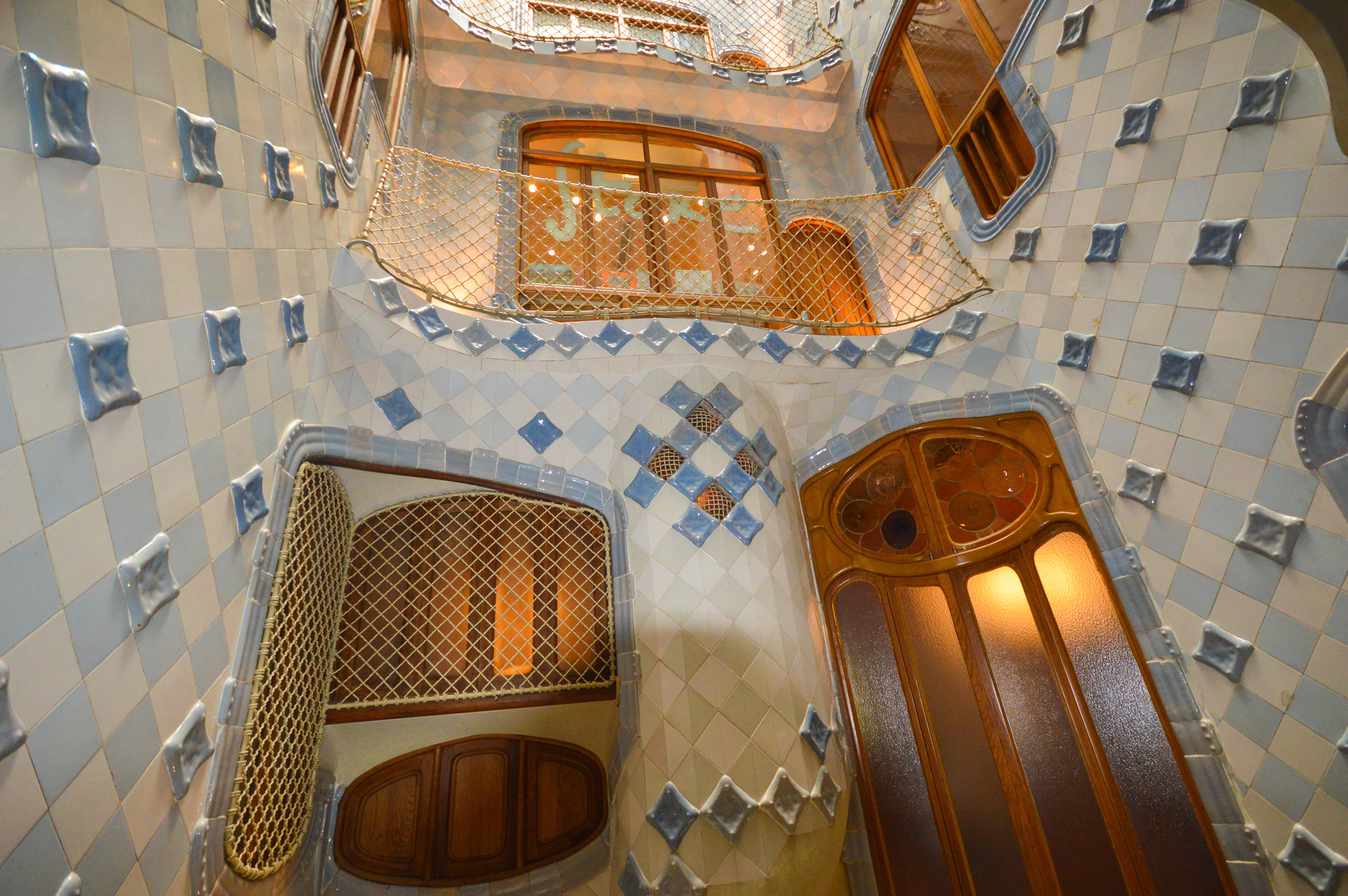The image depicts an interior staircase of a building adorned with intricate tile work. The walls are covered with light-colored tiles arranged in geometric patterns, accented with blue tiles. The staircase spirals upwards with wooden railings and doors, showcasing a blend of artistic and functional design elements.