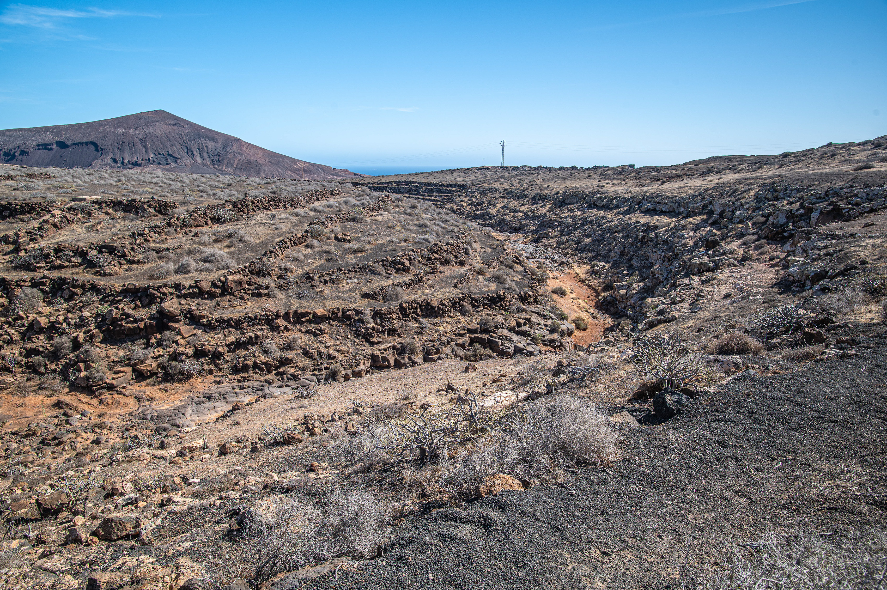 The image depicts a rugged, barren landscape with rocky terrain and sparse vegetation. In the background, there are hills or small mountains under a clear blue sky. A lone utility pole stands in the distance, indicating some human presence in this otherwise natural and desolate area.