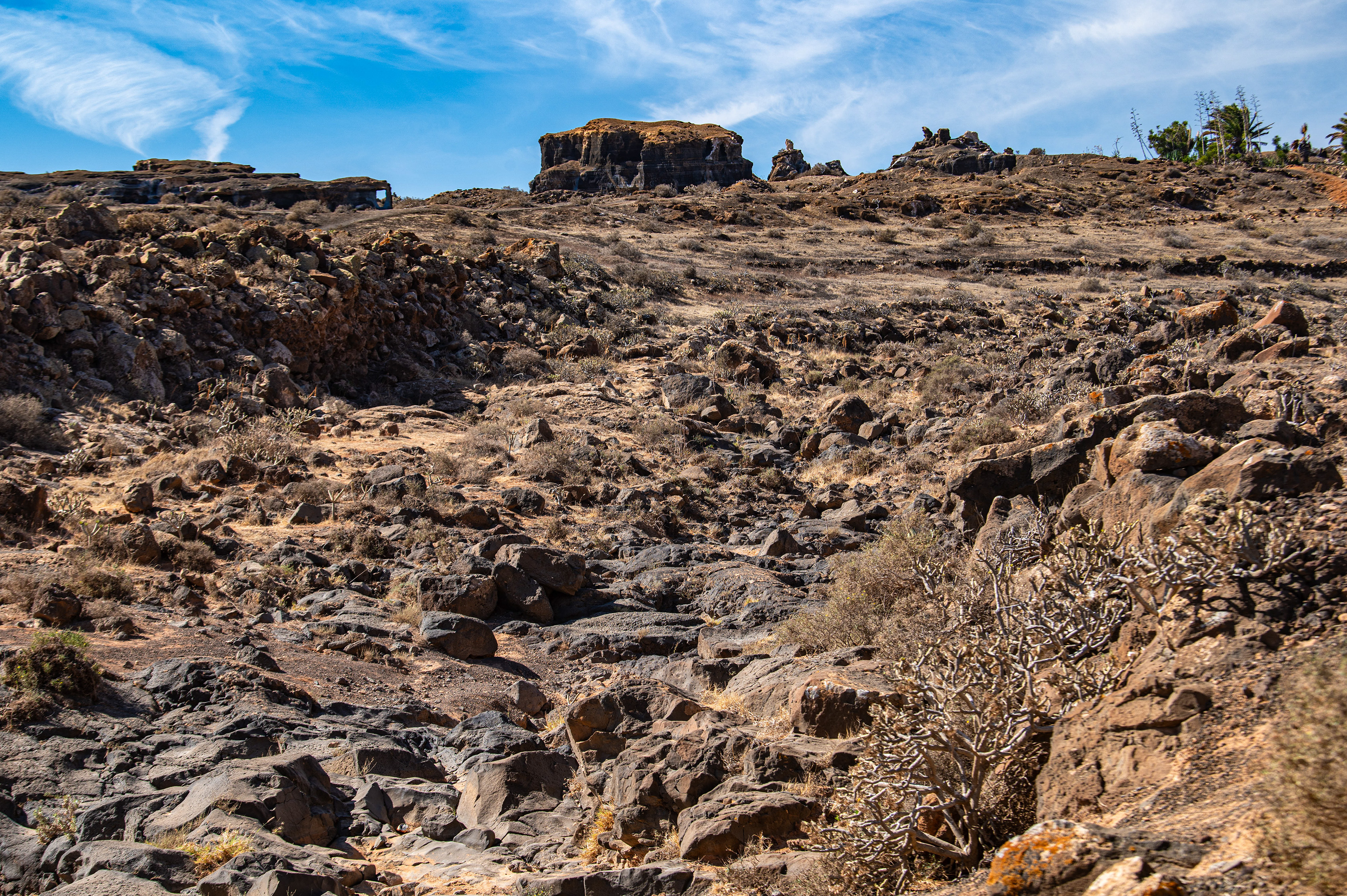 The image depicts a rugged, rocky landscape with a clear blue sky overhead. The terrain is characterized by large, jagged rocks and sparse vegetation, suggesting a harsh, arid environment. The scene is likely a natural, remote area with minimal human presence.