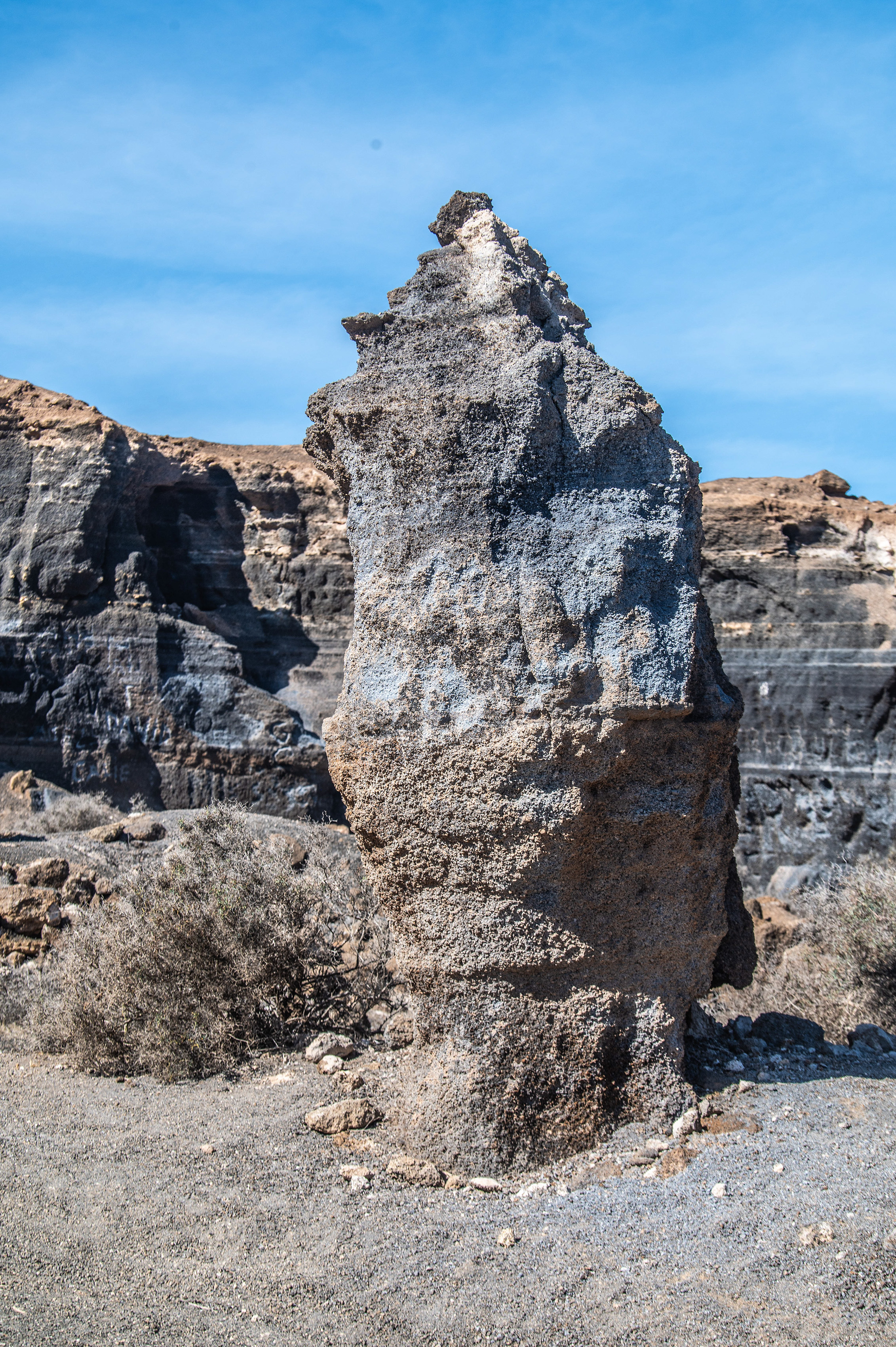 The image depicts a unique rock formation in a desert-like environment. The central feature is a tall, narrow rock pillar with a rough, textured surface, standing prominently against a clear blue sky. The surrounding area consists of rugged, rocky terrain with various rock formations and sparse vegetation. The overall scene suggests an arid, possibly volcanic landscape.
