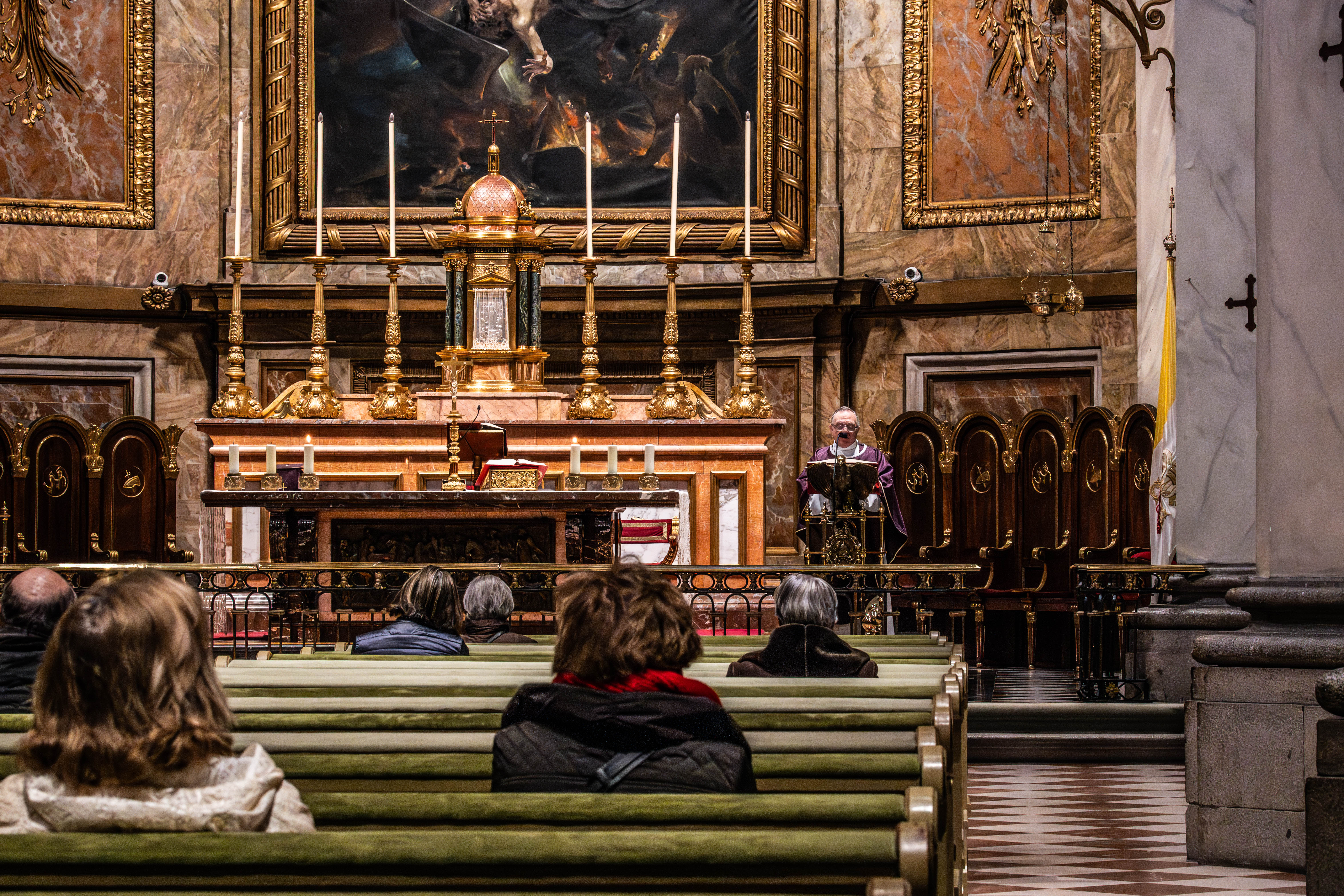 The image depicts an interior of a church where a religious service is taking place. The altar is elaborately decorated with candles, religious artifacts, and a large painting above it. A priest stands at a podium to the right of the altar, addressing a small congregation seated in the pews. The church features ornate wooden and marble elements, contributing to a solemn and reverent atmosphere.