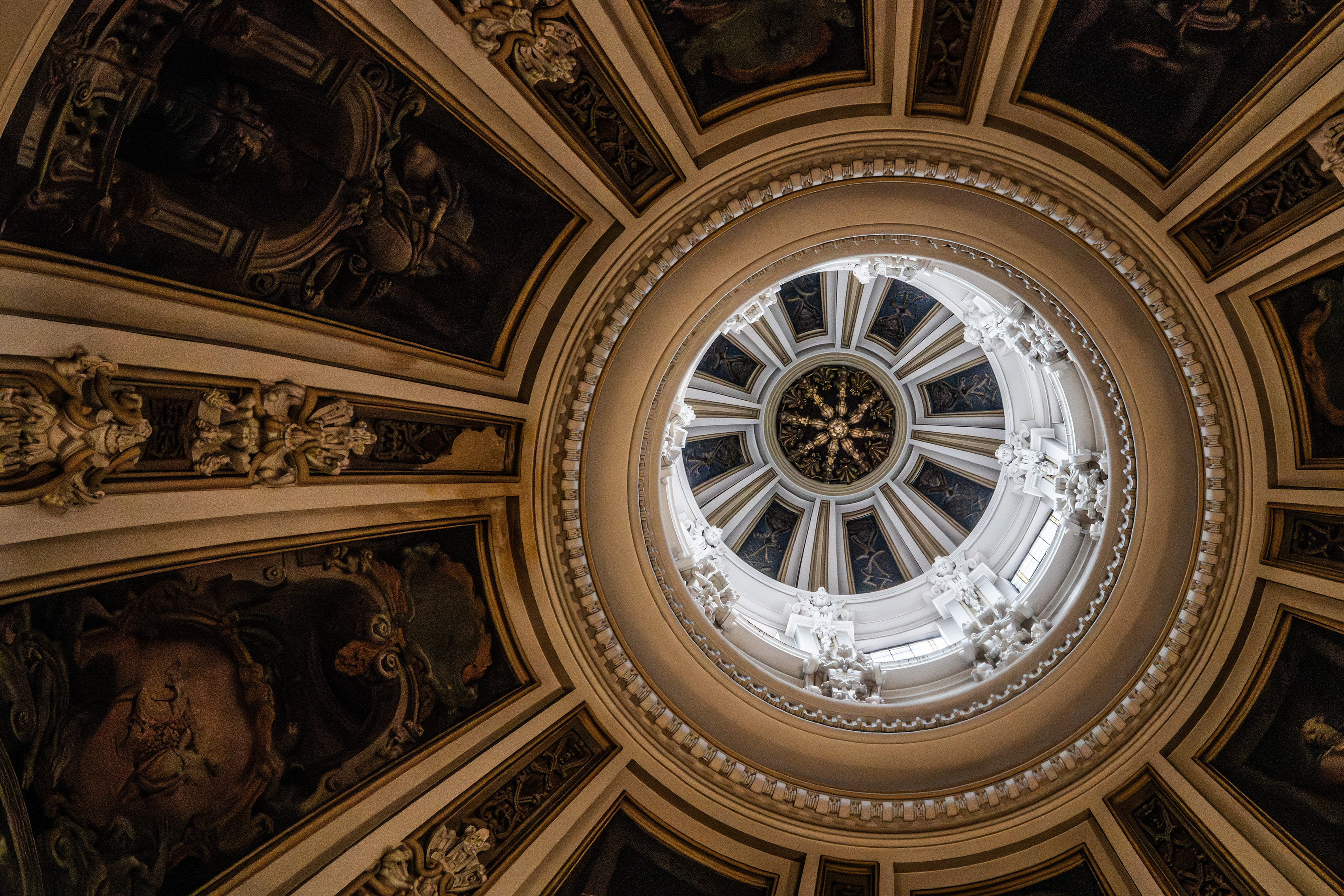 The image depicts an ornate ceiling with intricate artwork and architectural details, including frescoes, sculptures, and a central dome with a skylight.