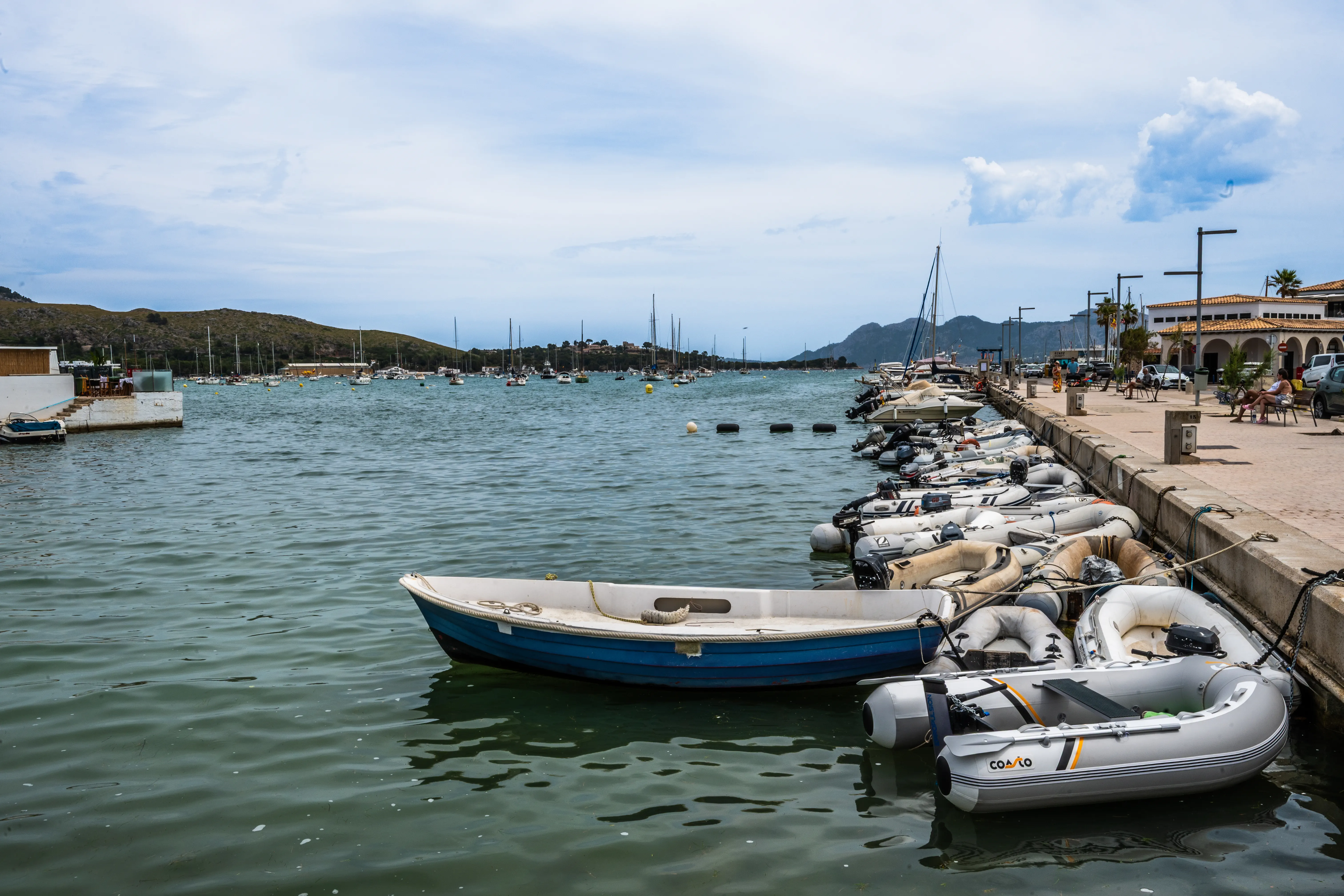 A marina with various boats, including a blue and white rowboat and several inflatable dinghies, docked along a promenade with buildings and hills in the background under a cloudy sky.