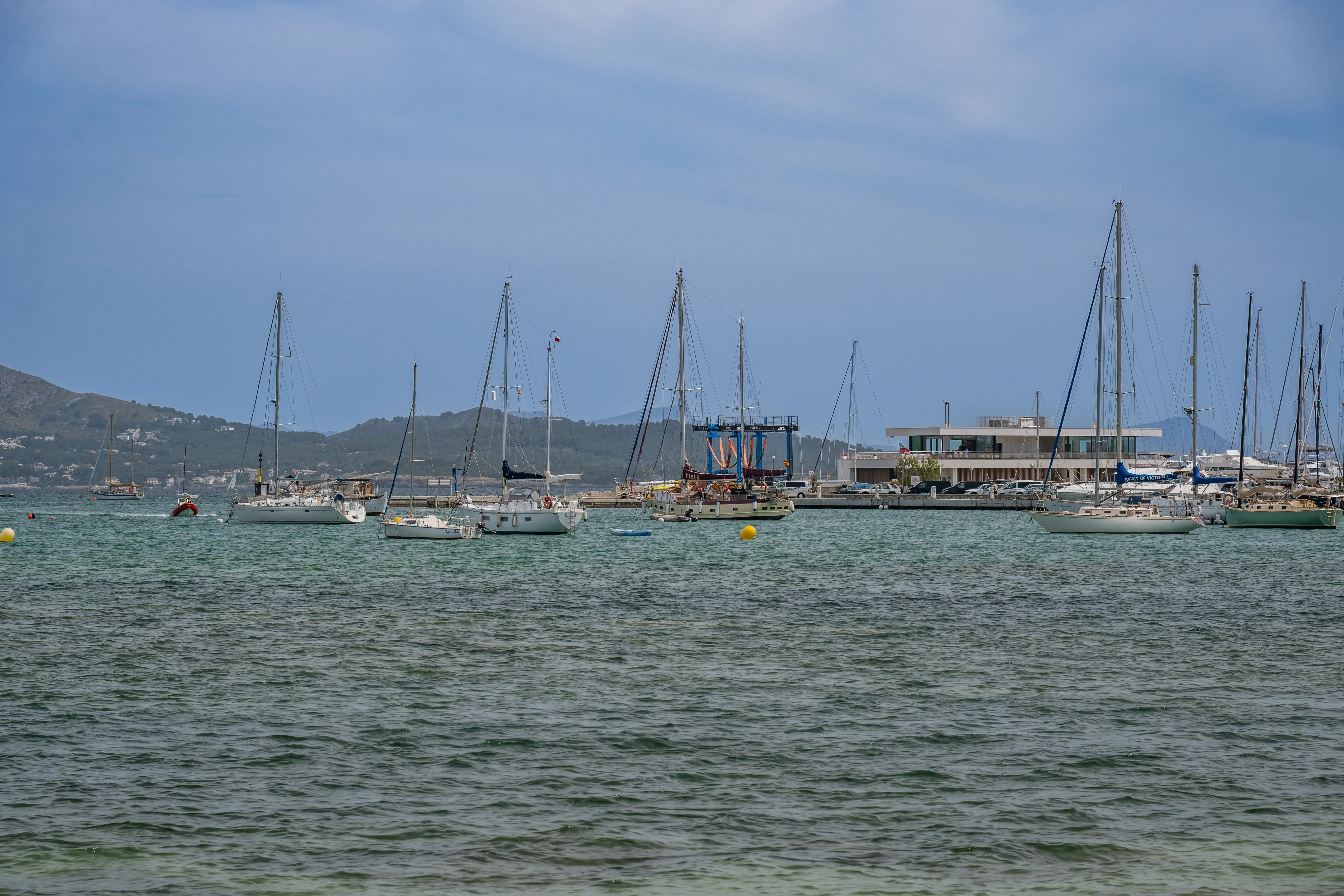 A wide shot of a marina harbour with many sailboats of different sizes moored in the water. In the background, there is a modern building and hills under a cloudy sky.