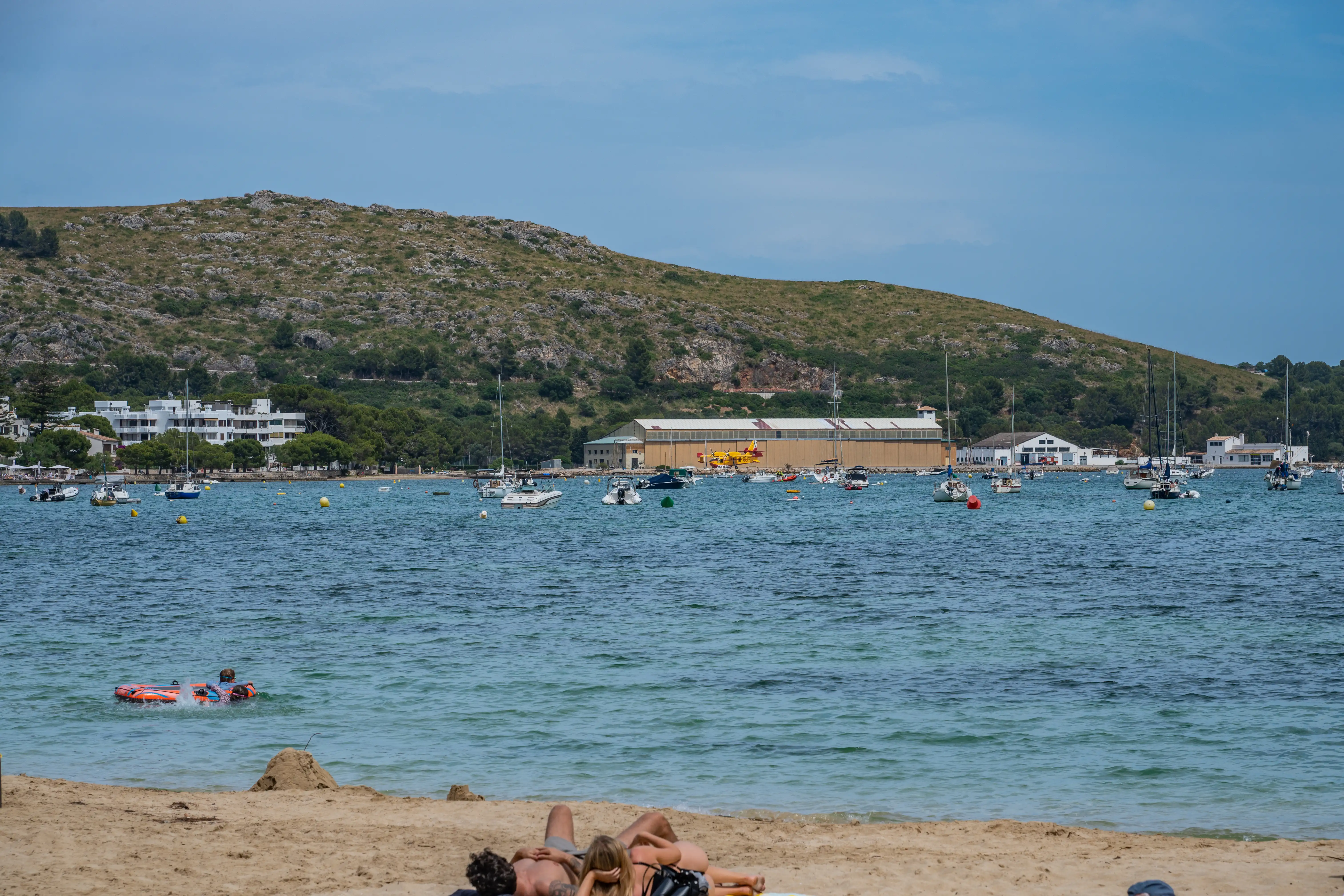 A bay with numerous boats at anchor, a sandy beach in the foreground with sunbathers, and a hillside with buildings and a yellow seaplane in the background under a clear sky.