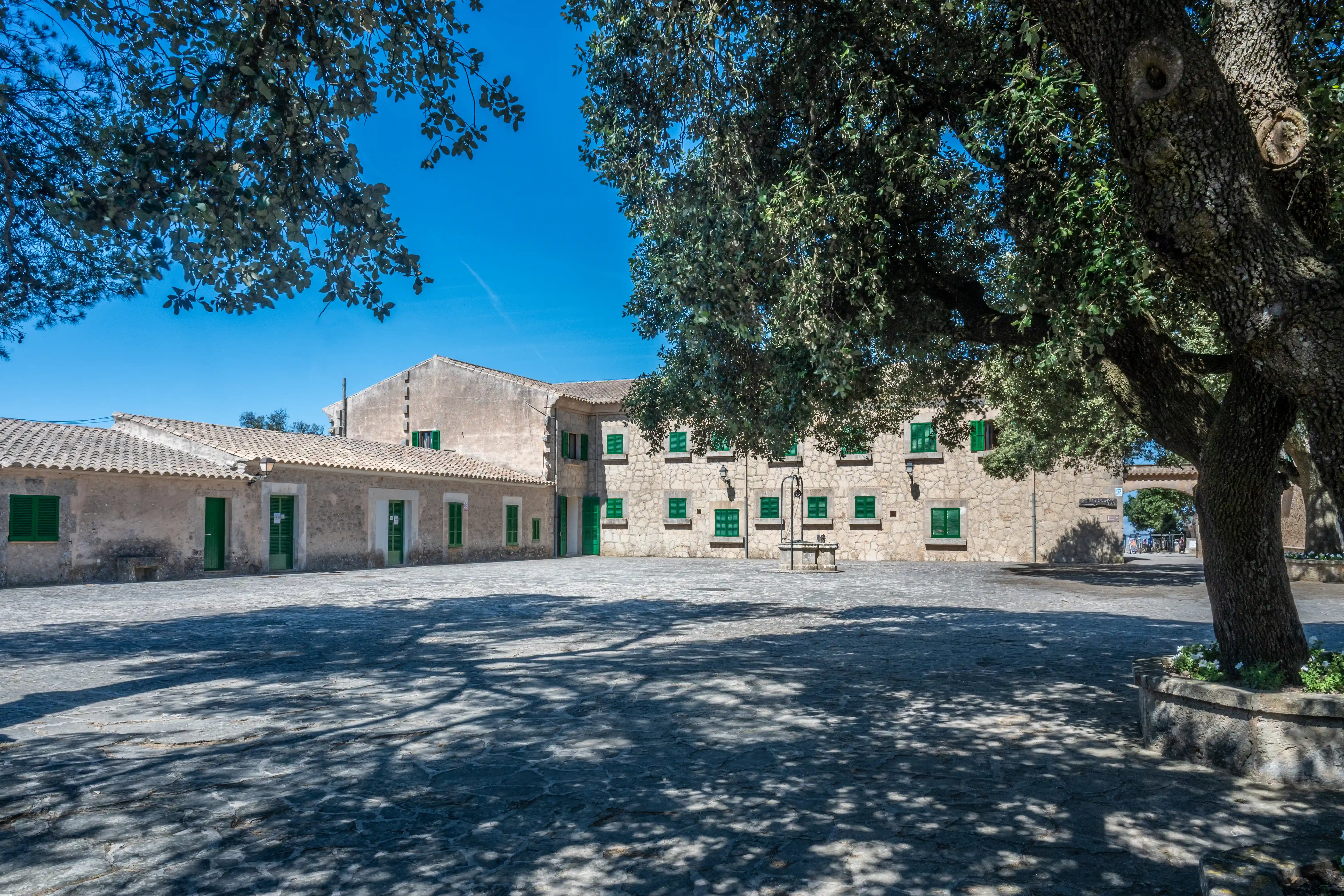 A sunny courtyard in front of a rustic stone building with a tiled roof and bright green shutters. Large trees cast shadows on the stone-paved ground. A stone well is visible in the centre.