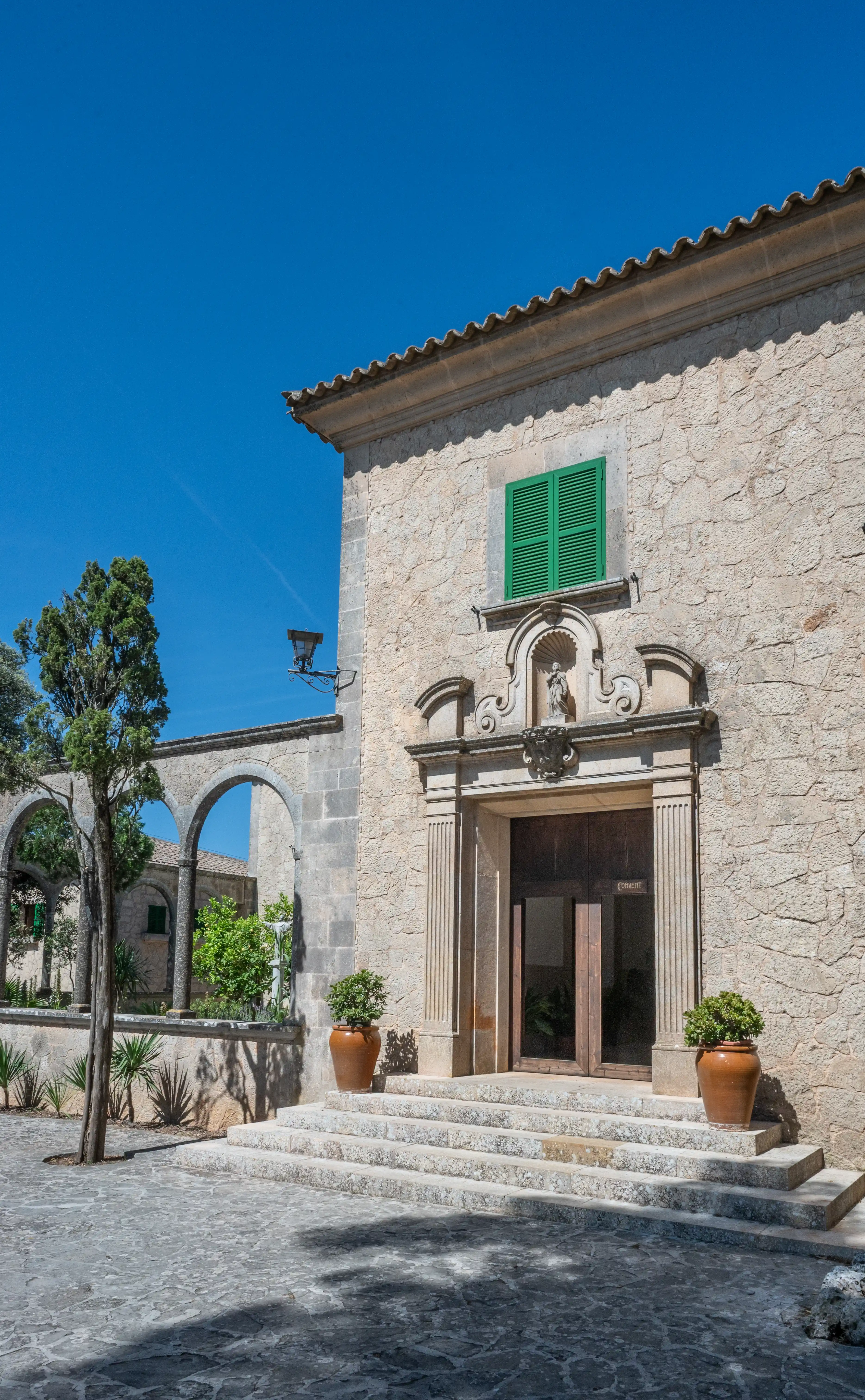 A sunlit view of a traditional stone building with a prominent entrance featuring wooden doors, ornate carvings, and a vibrant green shuttered window. Stone arches and a tall tree are visible to the left, with potted plants decorating the stone steps leading to the entrance.