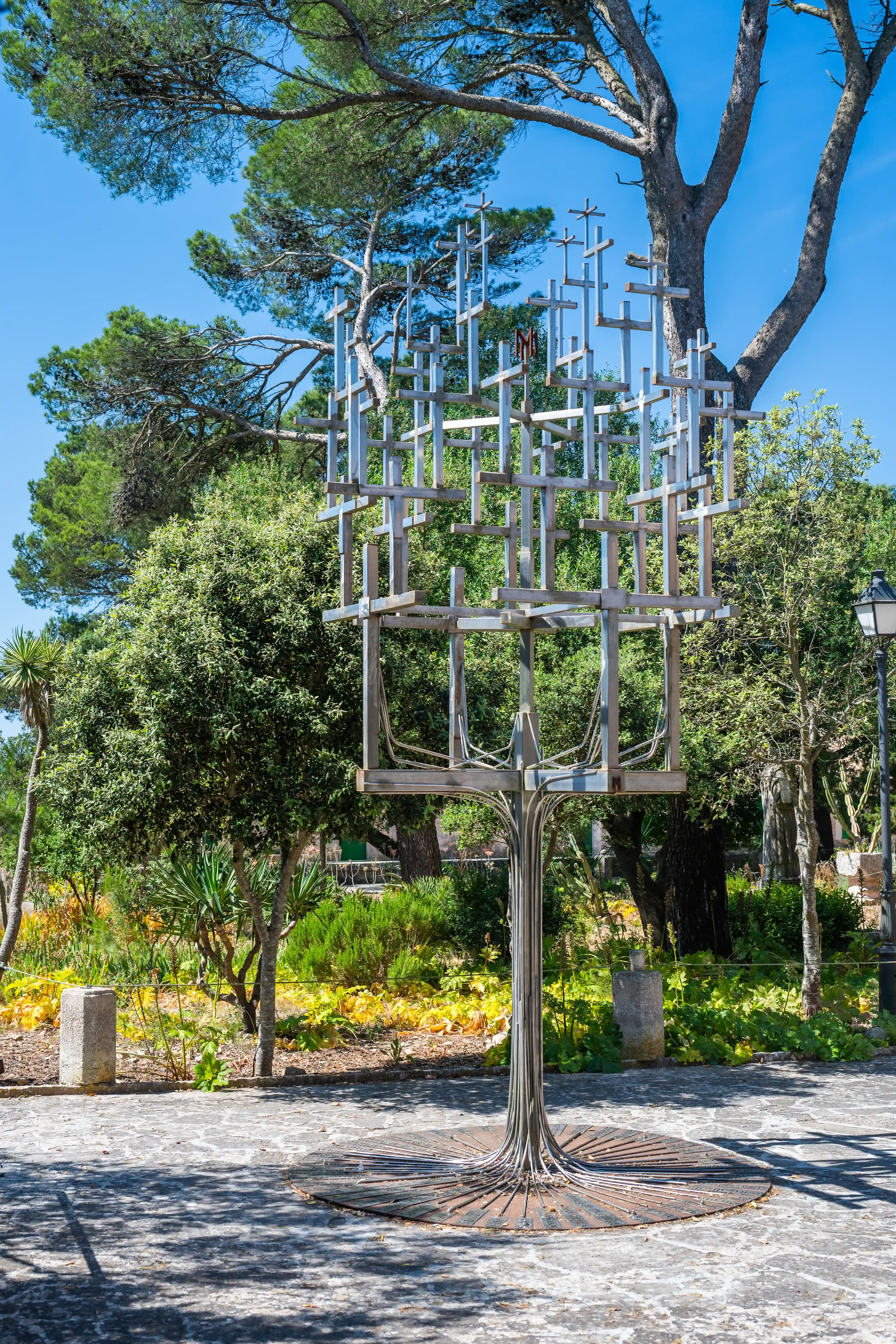 A contemporary sculpture made of crisscrossed metal bars, resembling a stylised tree or abstract form, is displayed outdoors in a verdant garden on a sunny day.