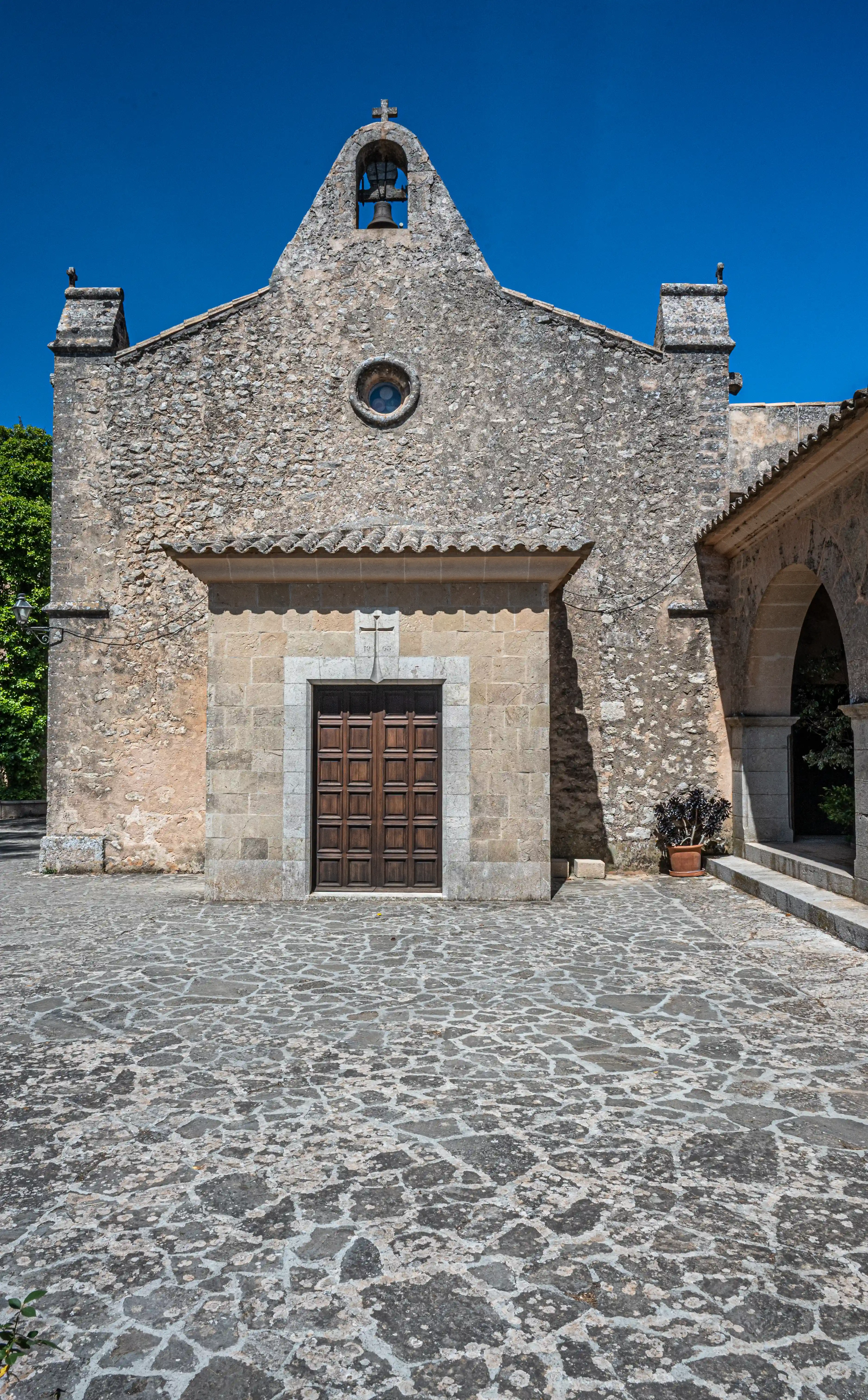 Front facade of a rustic stone chapel featuring a bell tower with a cross, a circular window, a wooden double door, and a tiled roof. A cobblestone courtyard extends to the foreground, and an arched walkway is visible to the right.