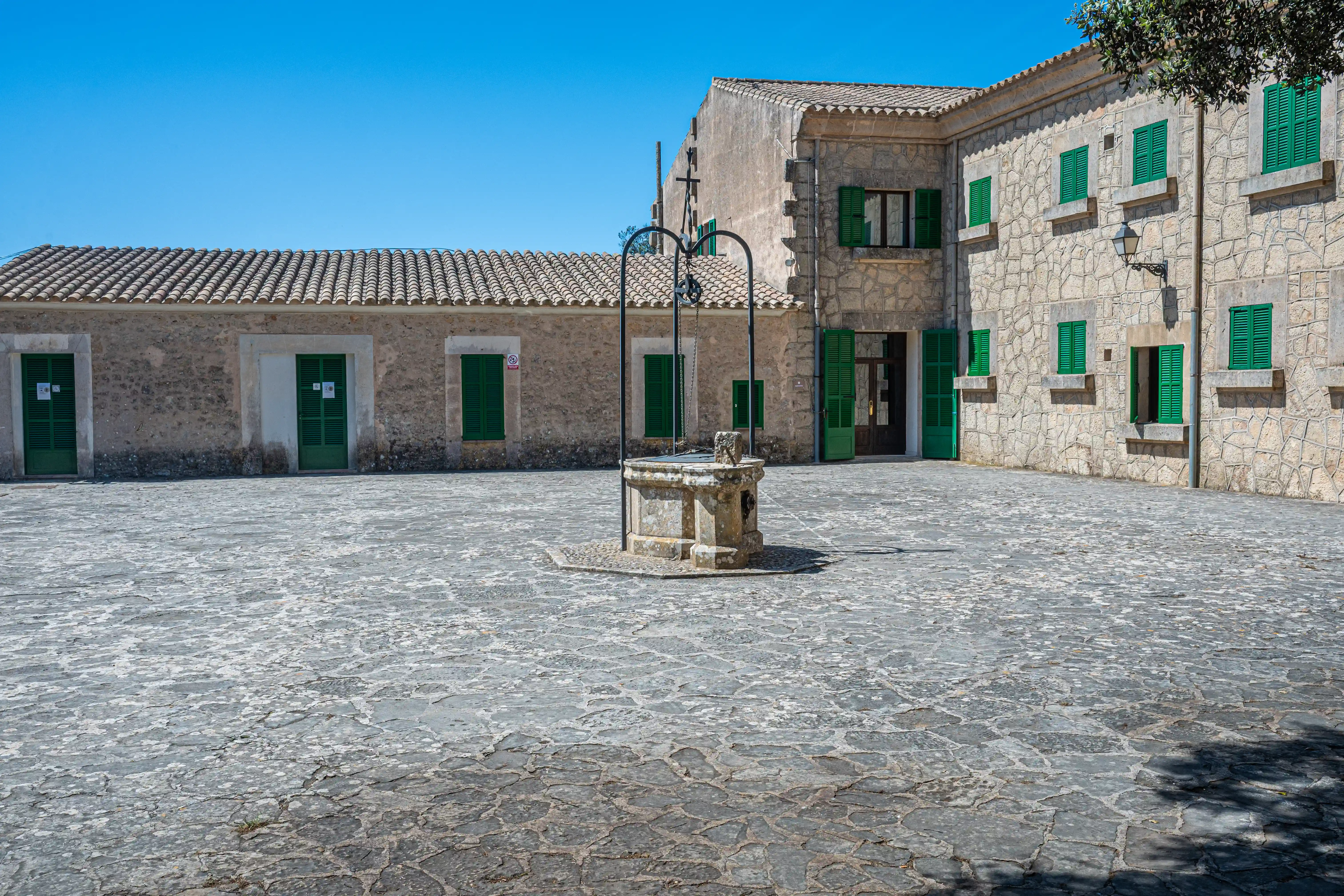 An image of a stone courtyard with a central well and traditional stone buildings with green shutters.