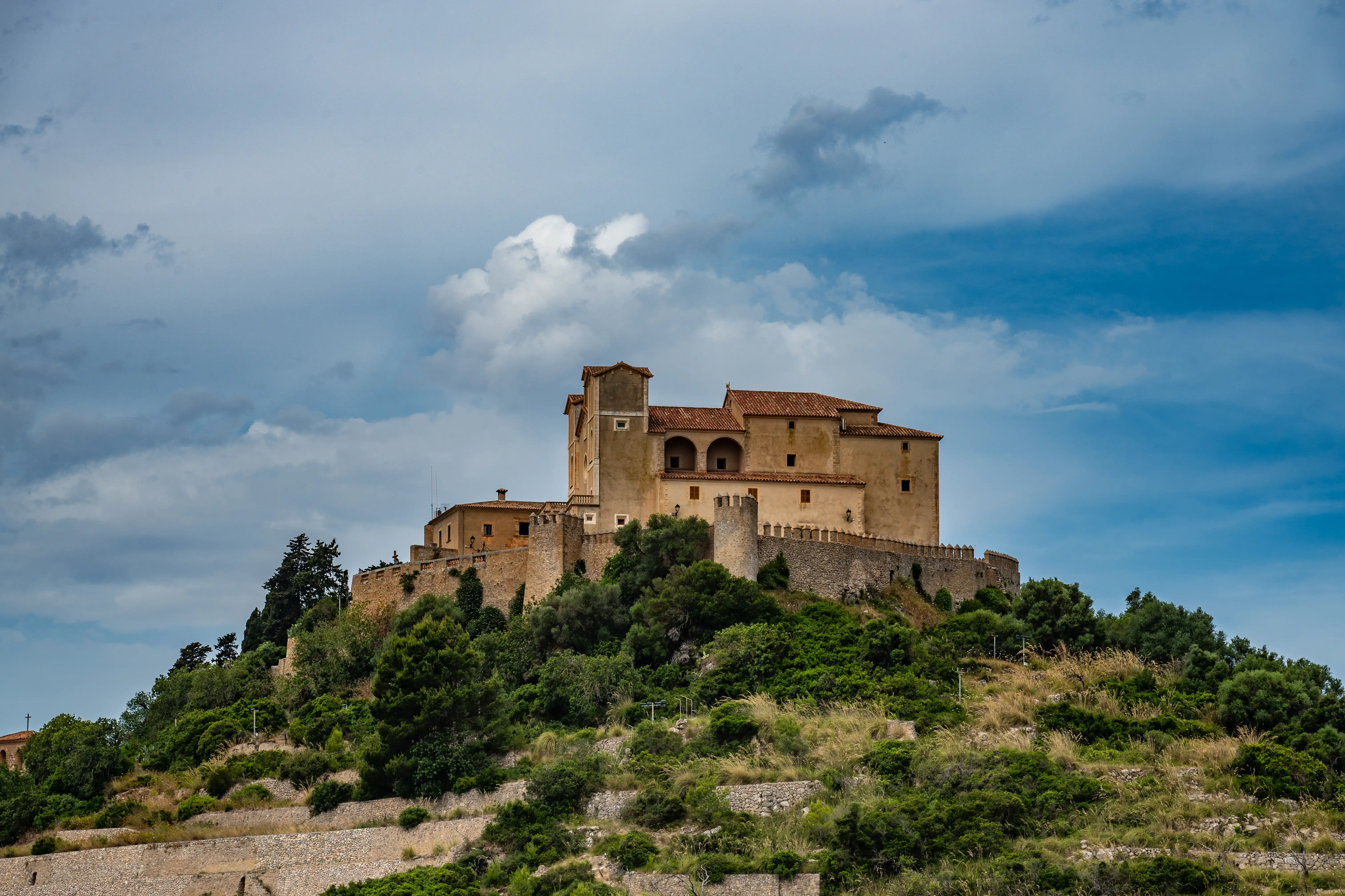 A wide-angle shot of a stone castle complex with terracotta roofs, built on a densely vegetated hillside, under a partly cloudy blue sky.