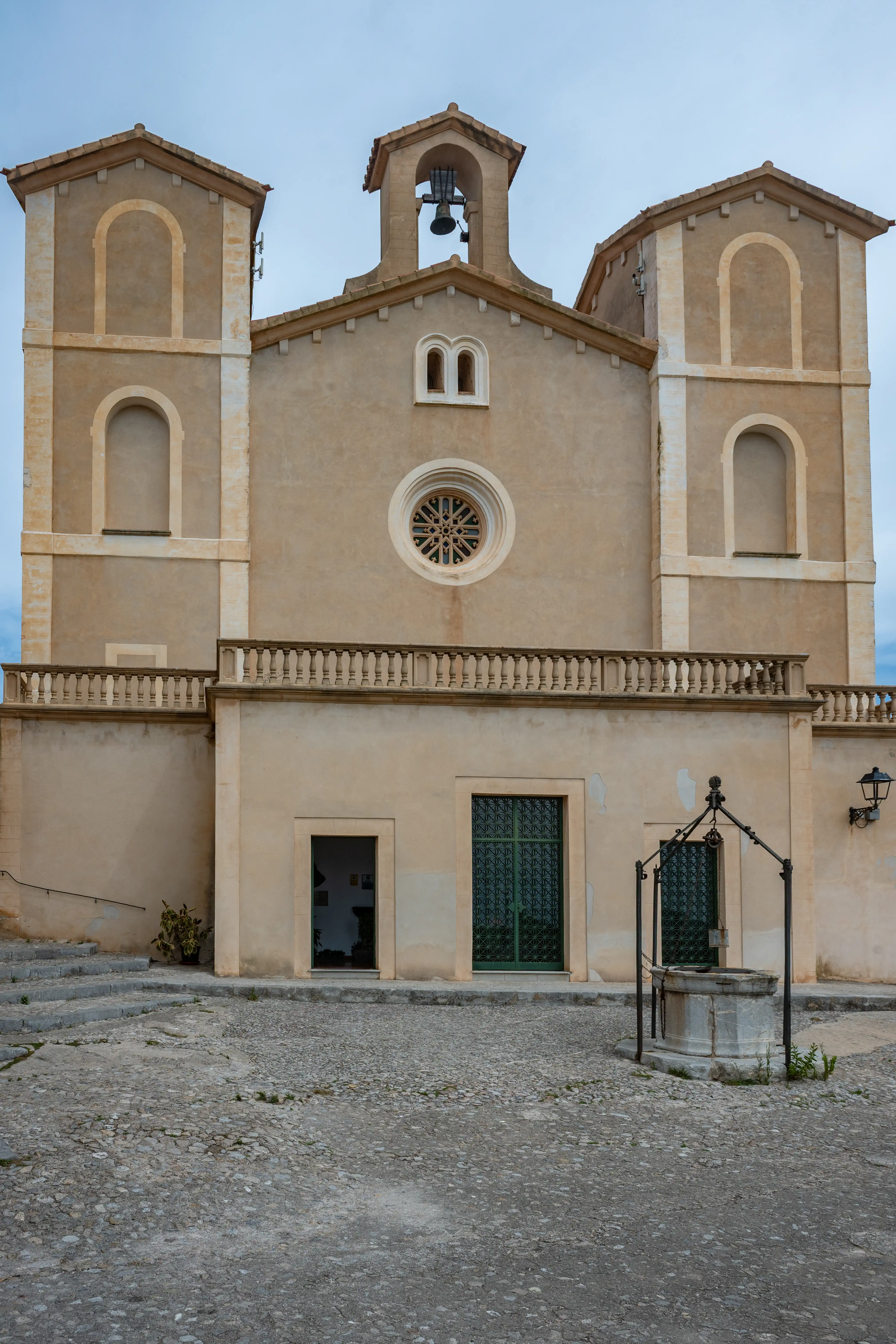 Exterior view of a religious building with a central bell tower and two side towers, a balcony, a circular decorative window, and an old stone well in a cobbled plaza.