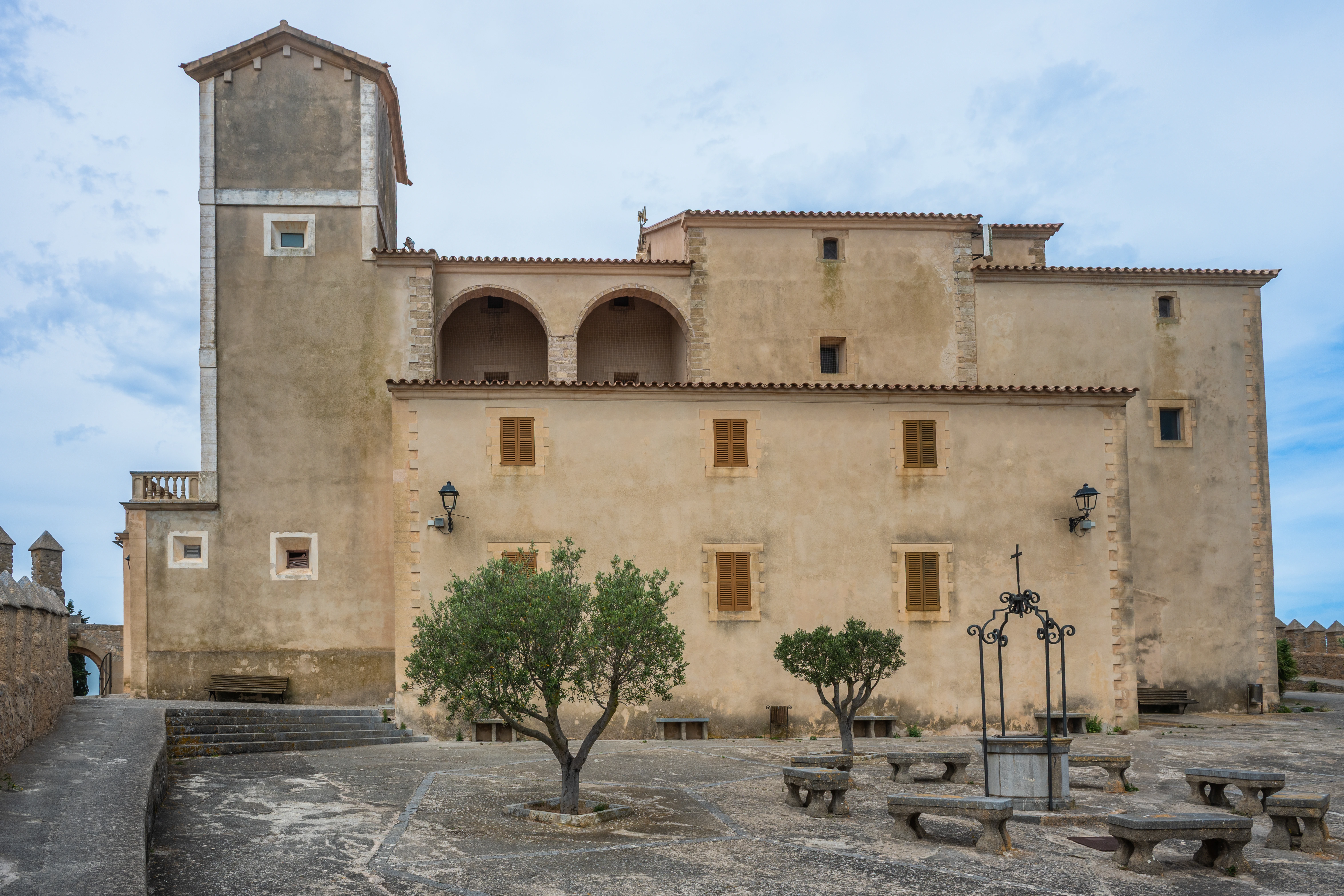 A view of a historic building with a tower and arched colonnade, situated in a courtyard with stone benches, a well, and olive trees under a cloudy sky.