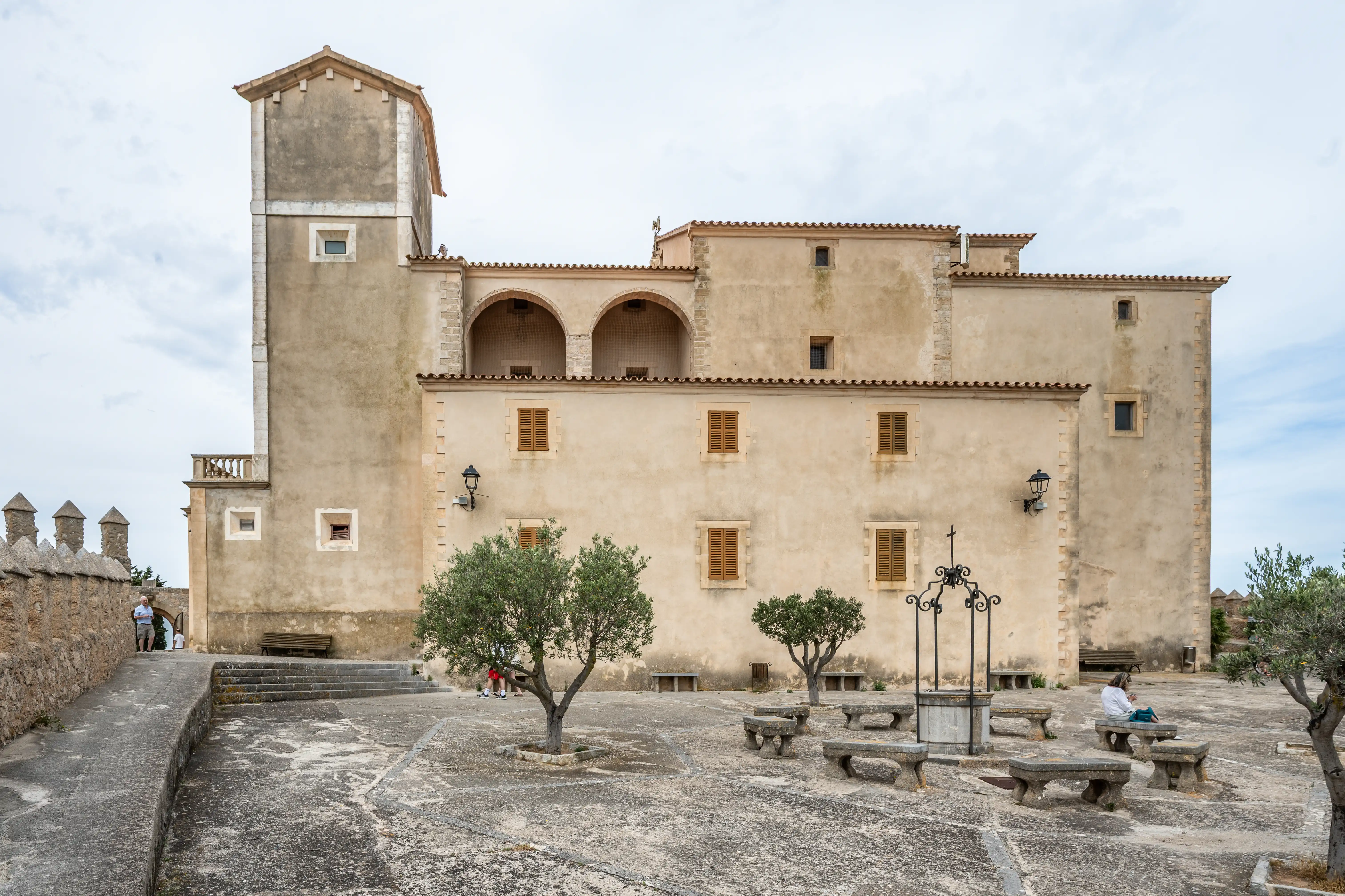 A stone courtyard in front of an old building with a tower, featuring stone benches, olive trees, and a well. A stone wall is to the left.