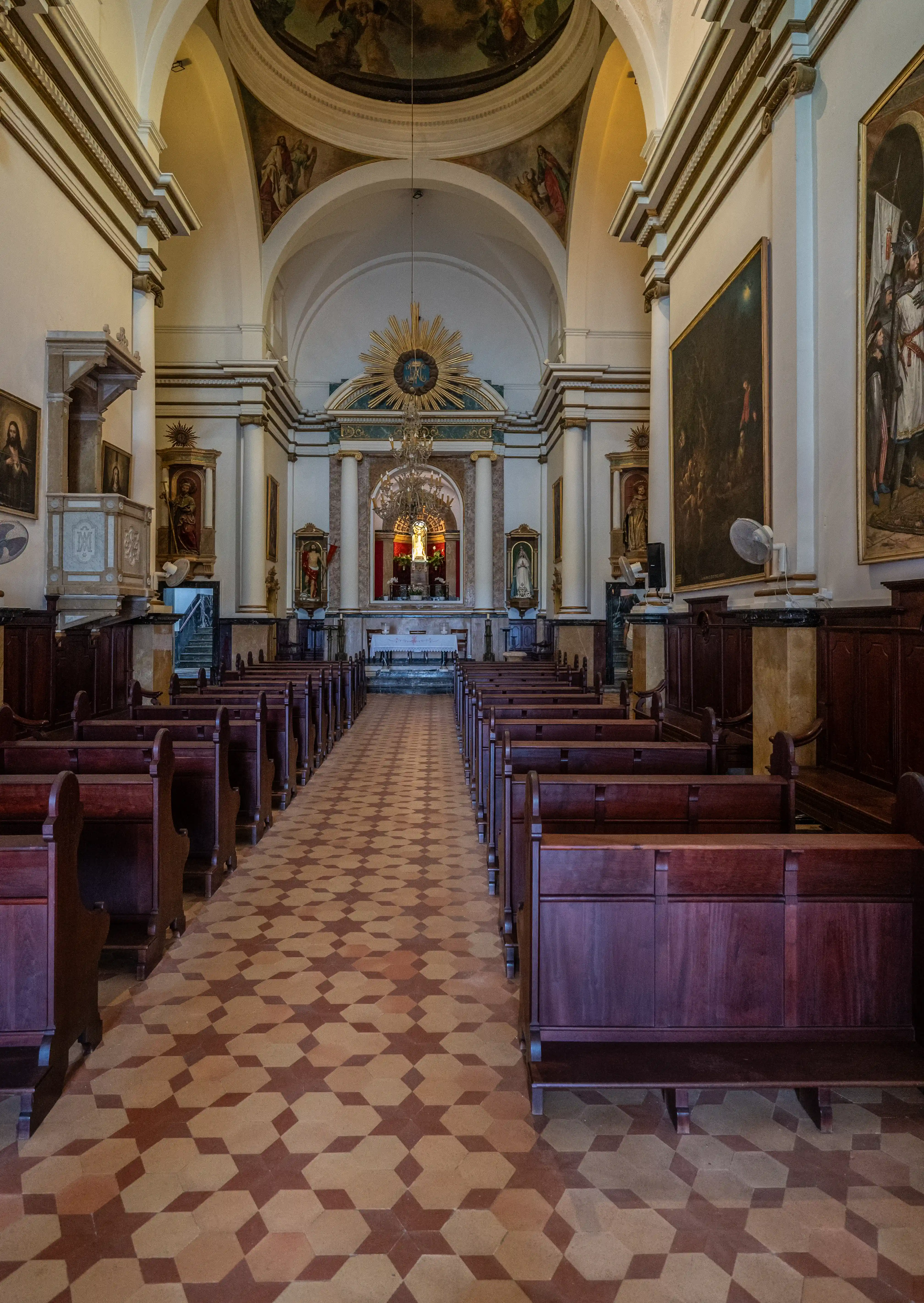 Interior of a church showing rows of dark wooden pews leading to an altar, with patterned tile flooring and religious artwork on the walls and ceiling.