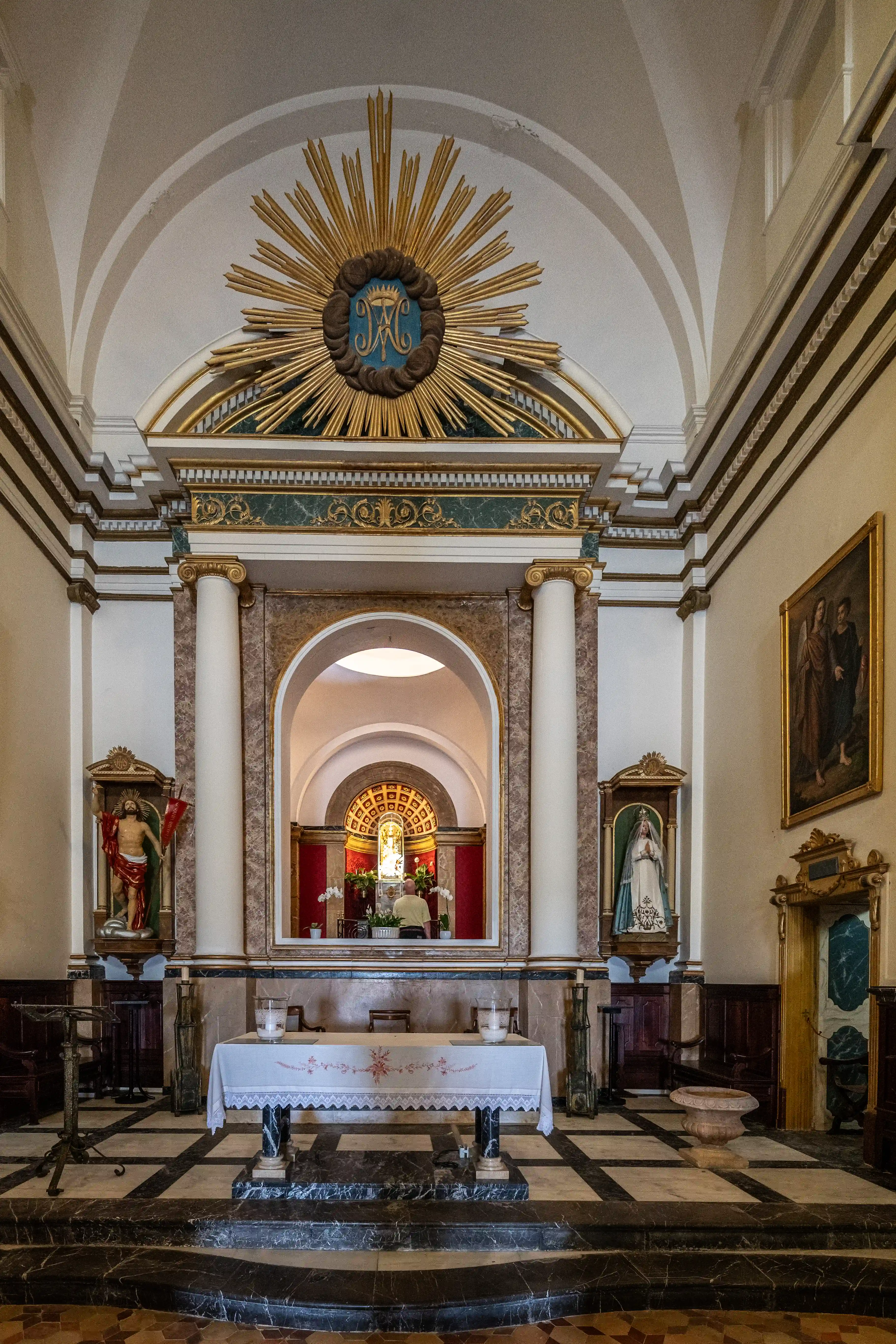 The main altar of a church, viewed from the nave. The altar is covered with a white cloth. To the left is a statue of Jesus, and to the right is a statue of the Virgin Mary. Above the altar is a sunburst motif with a monogram. In the background, a person is attending to the area behind the altar.