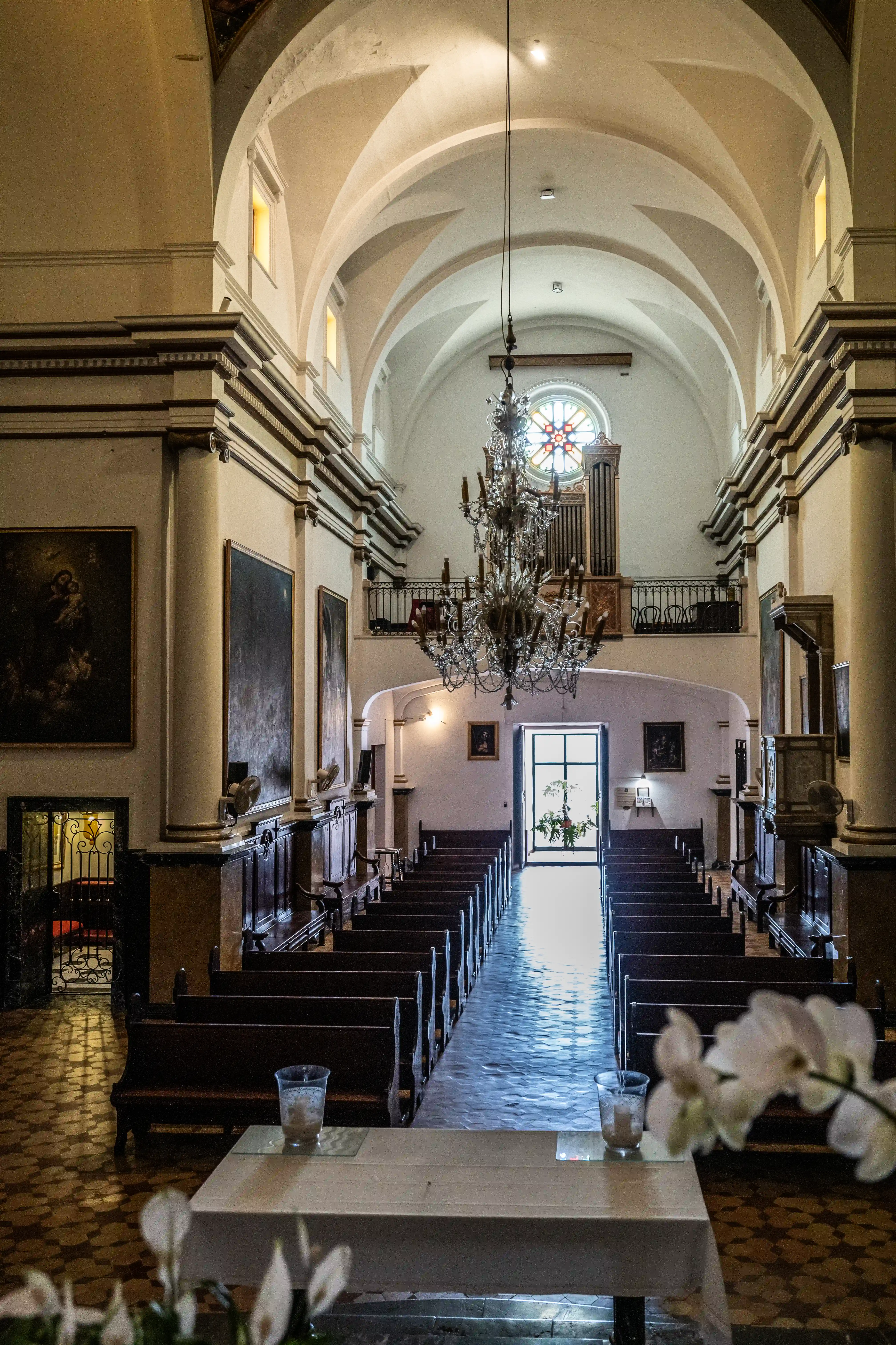  Interior view of a church nave, looking towards the altar and entrance, featuring pews, a large chandelier, vaulted ceiling, pipe organ, and stained-glass window.