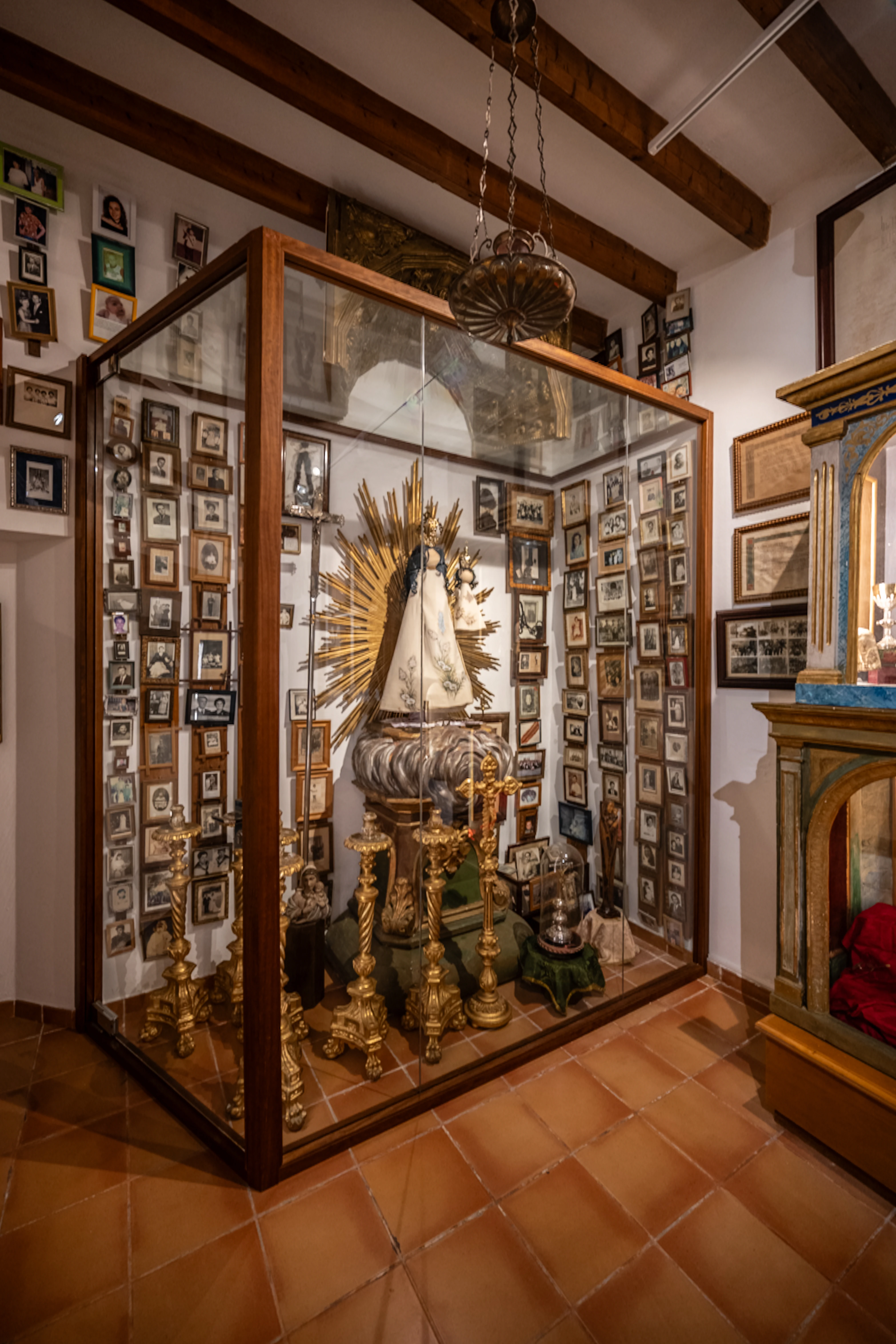 A glass display case filled with a religious statue and surrounded by framed photographs, set against a white wall with wooden ceiling beams and a terracotta tiled floor.