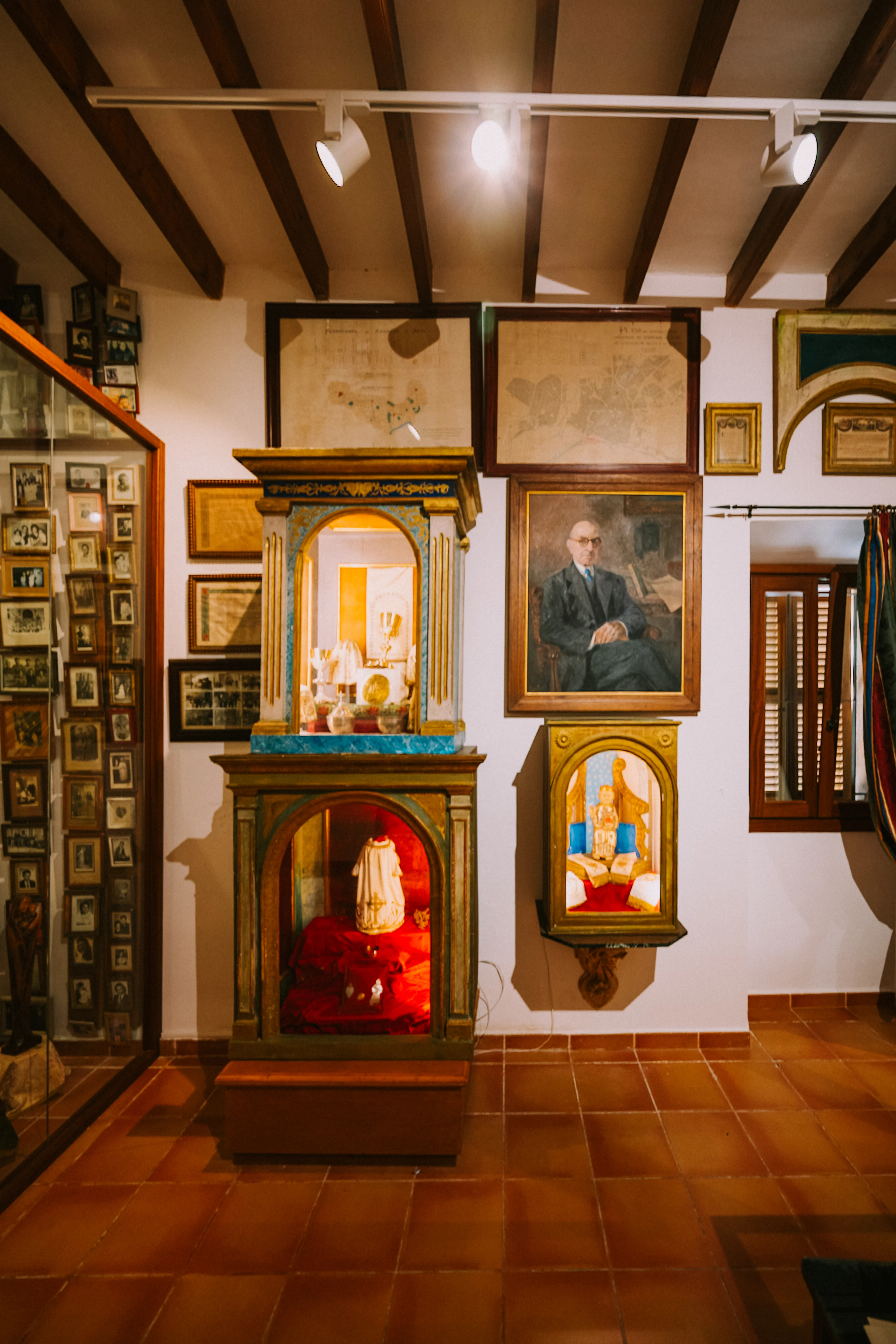  A museum display featuring a large framed portrait of a man in a suit, flanked by framed maps and smaller framed pictures. In the foreground, two intricately decorated wooden cabinets display religious figurines and vestments, with a glass display case filled with numerous small framed photographs to the left.
