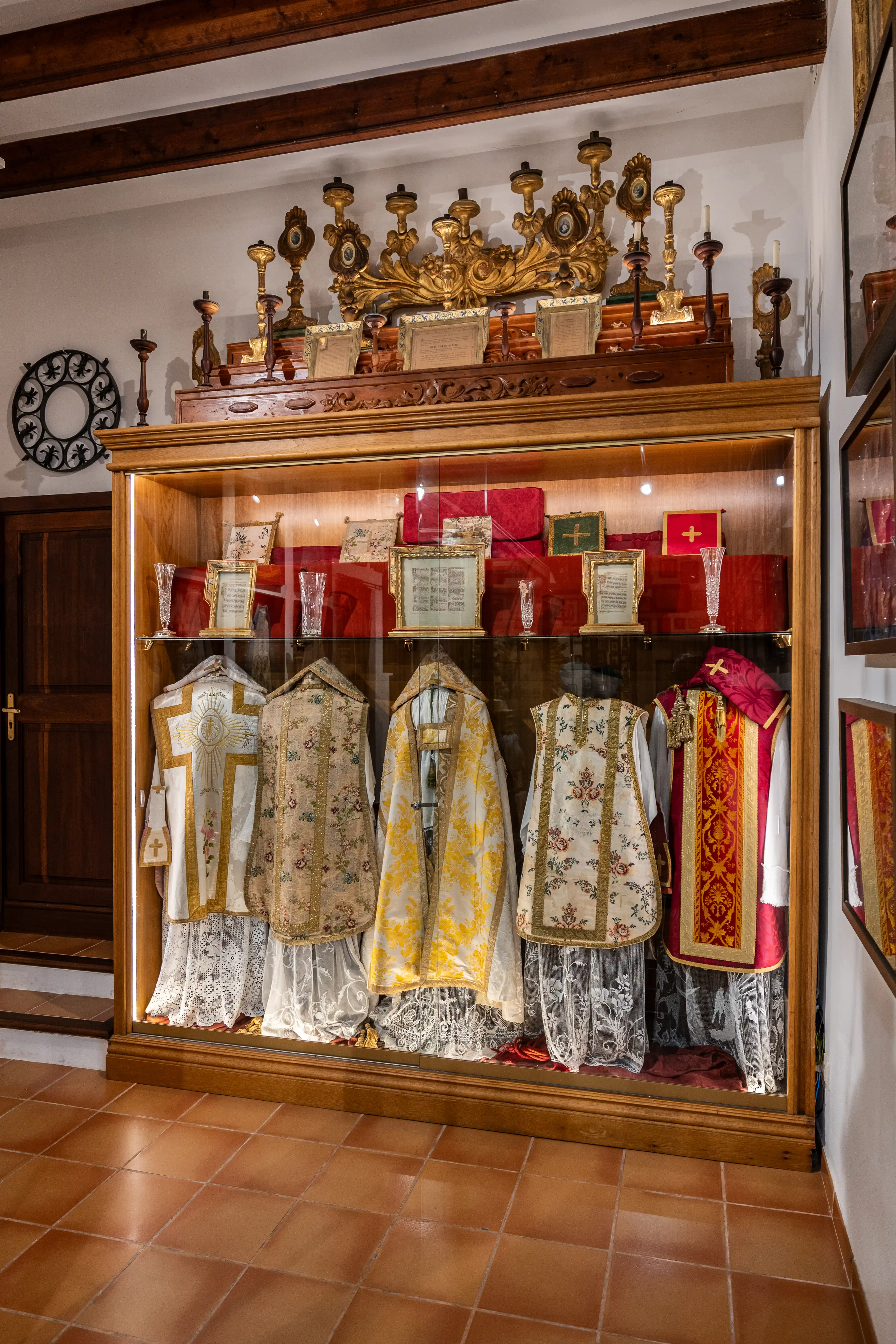 A wooden display cabinet filled with religious vestments and artefacts, including ornately embroidered robes, framed documents, and golden candelabras, illuminated for display.