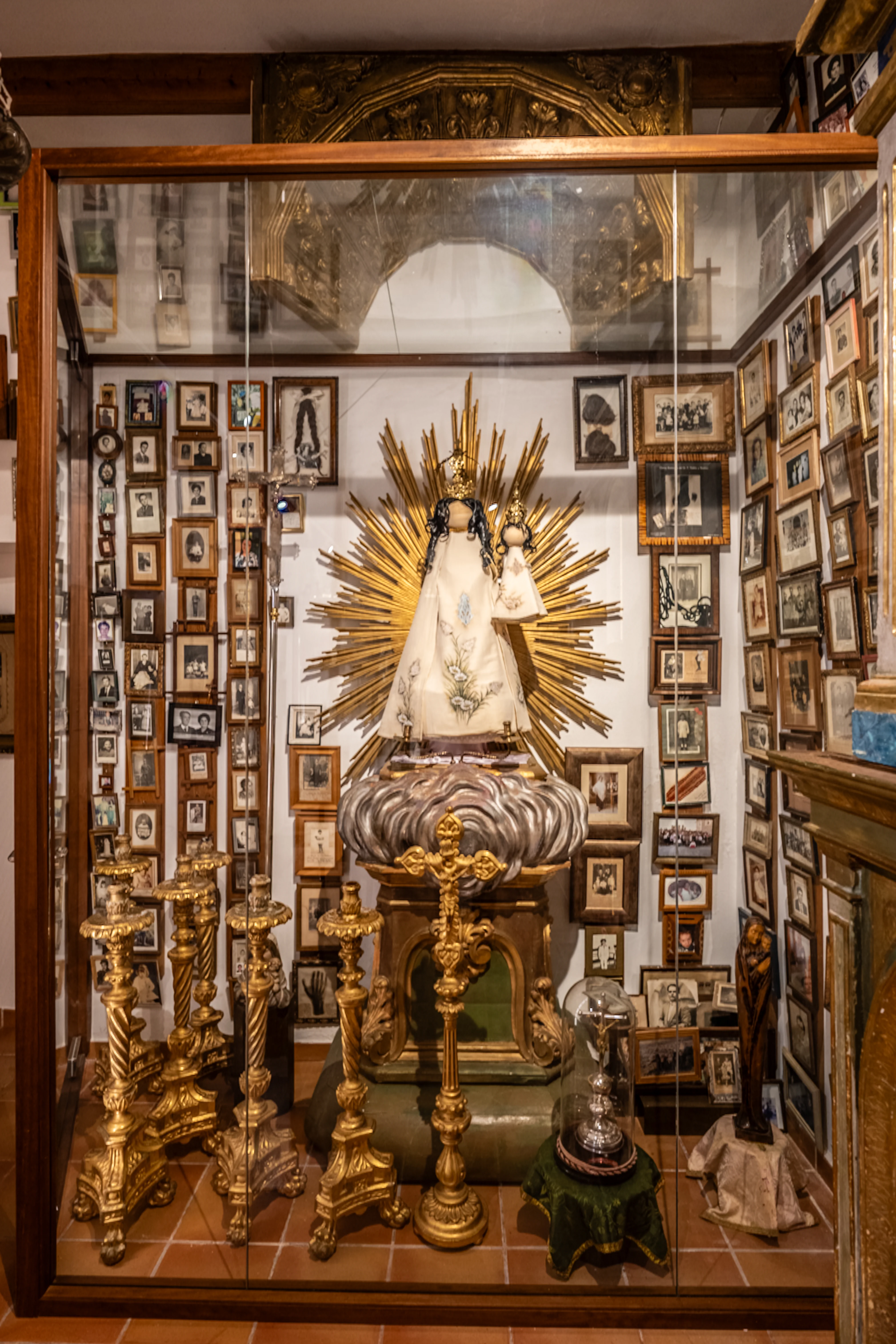 A glass display case exhibits a statue of the Virgin Mary and child radiating golden rays, flanked by elaborate golden candelabra and surrounded by a wall covered in an assortment of framed photographs.