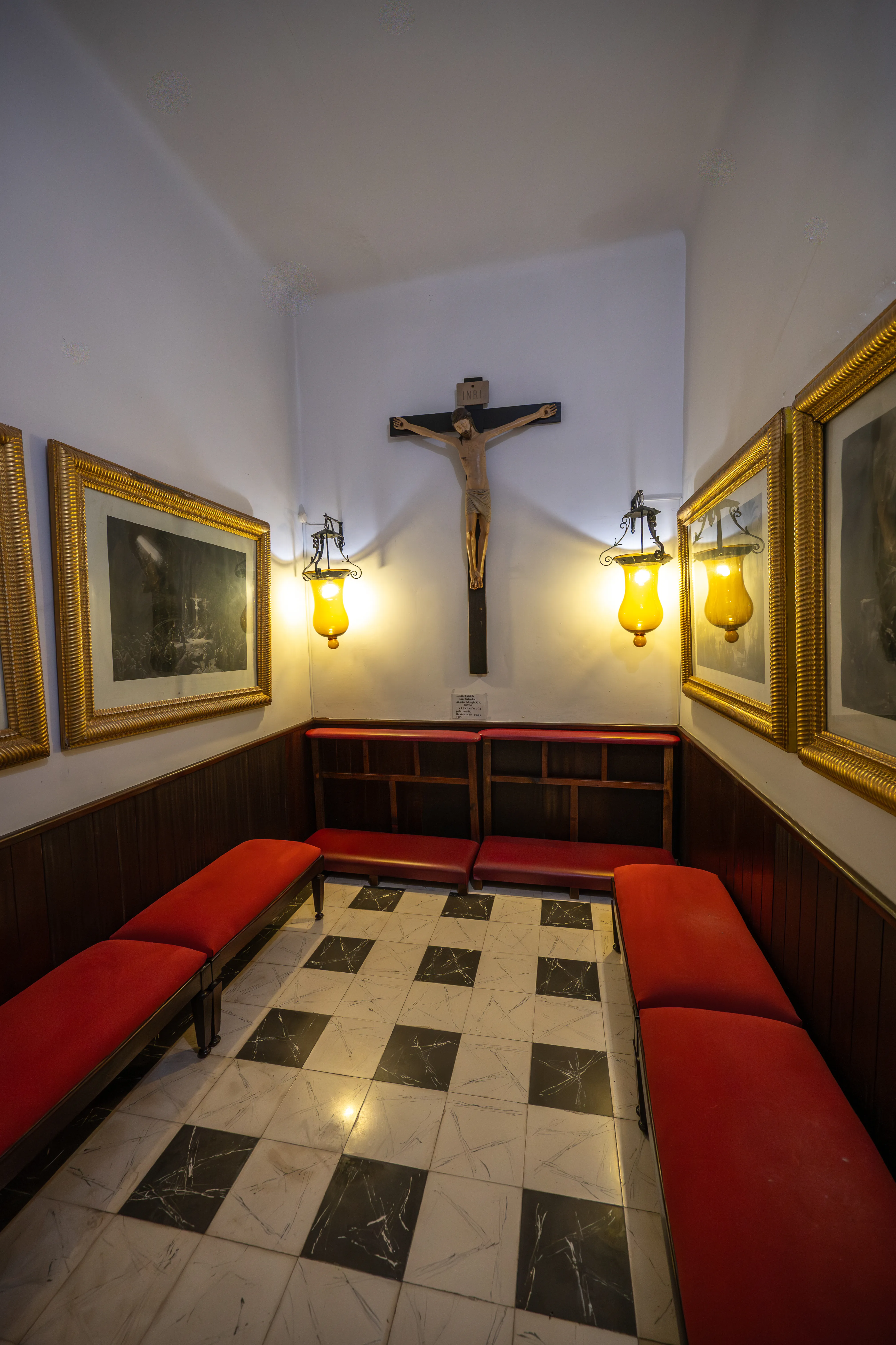  A religious room featuring a crucifix on the wall, illuminated by yellow lanterns, with red benches arranged along the sides and a checkered floor.