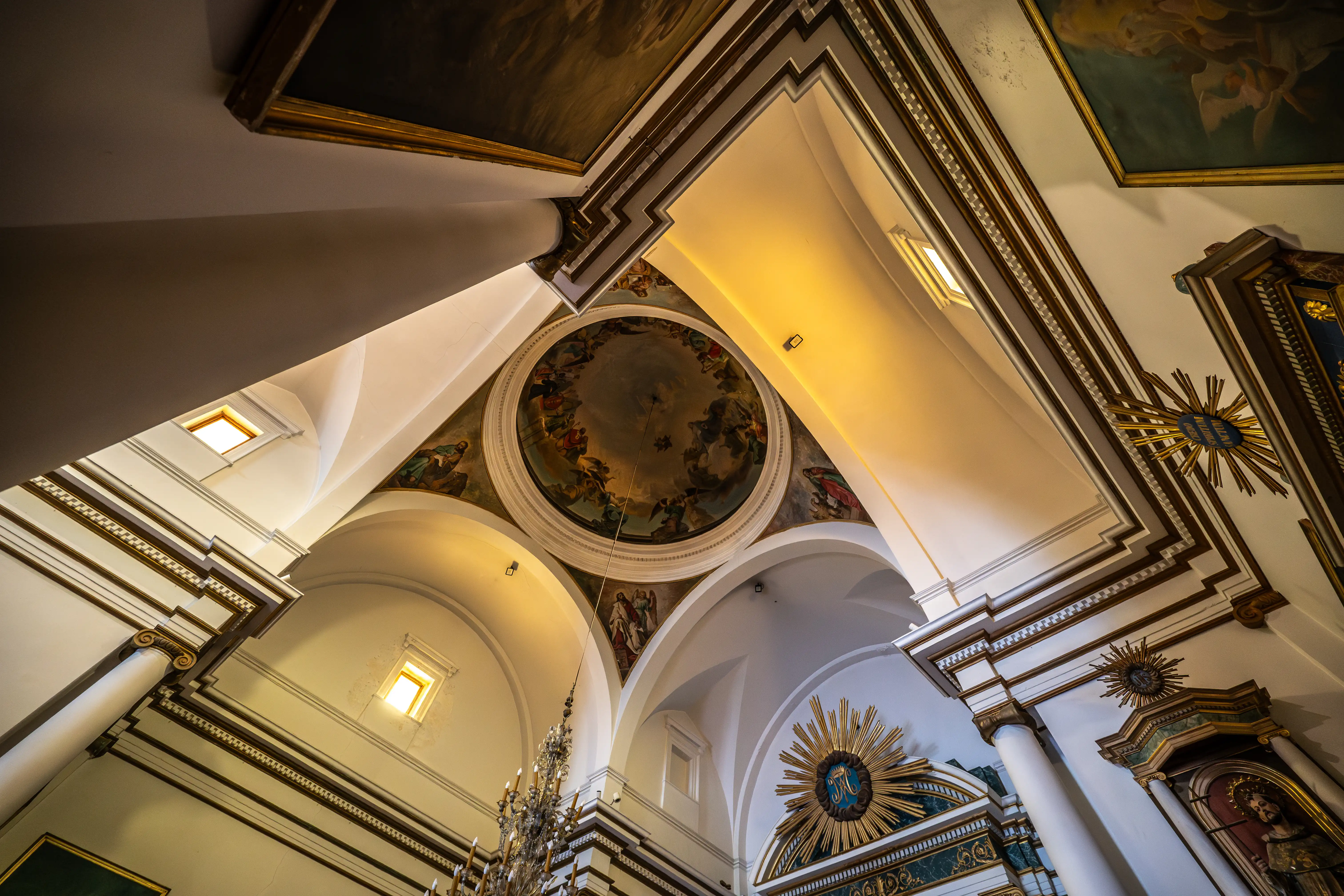 Interior view of a church ceiling with ornate frescoes, vaulted arches, golden trim, and a chandelier.