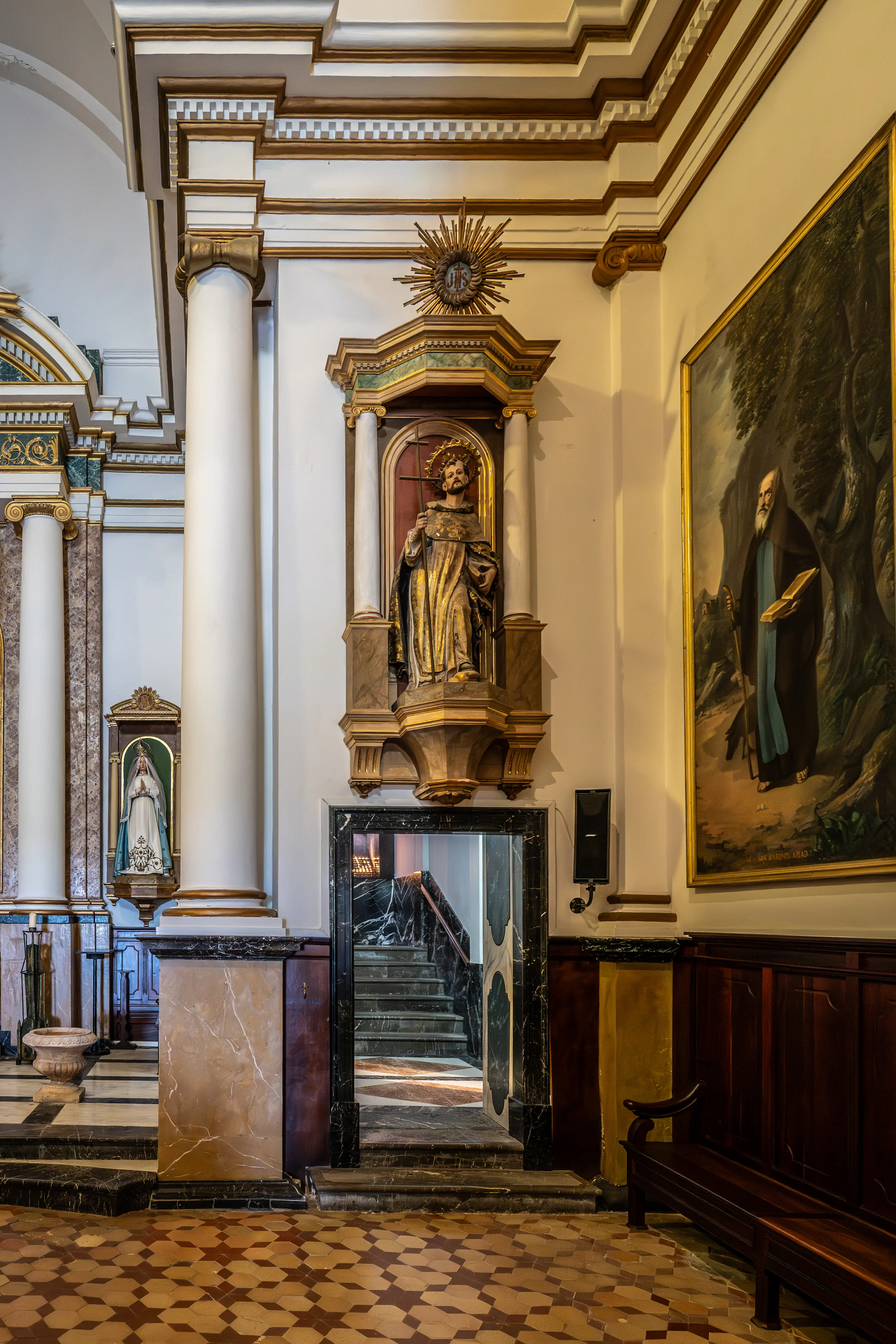 Inside a church, a golden statue of a saint with a halo stands prominently above an ornate niche. To the left, a smaller statue of the Virgin Mary is visible. A large painting of a robed man hangs on the right wall. A dark marble doorway leads to a staircase. The floor is tiled with a geometric pattern.