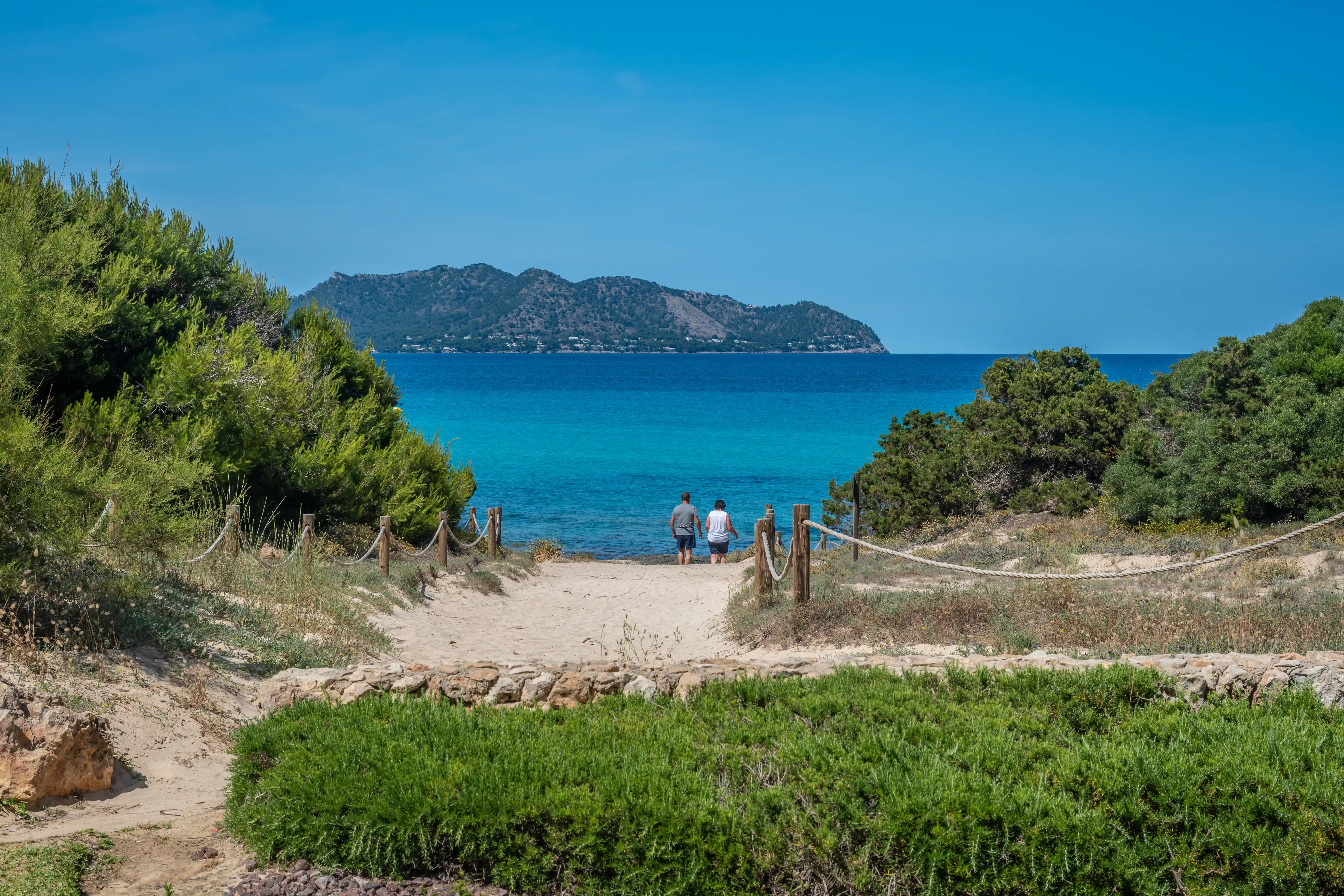 A sandy path through coastal vegetation leads to a clear turquoise sea, with a mountainous island in the background and two people walking towards the water.