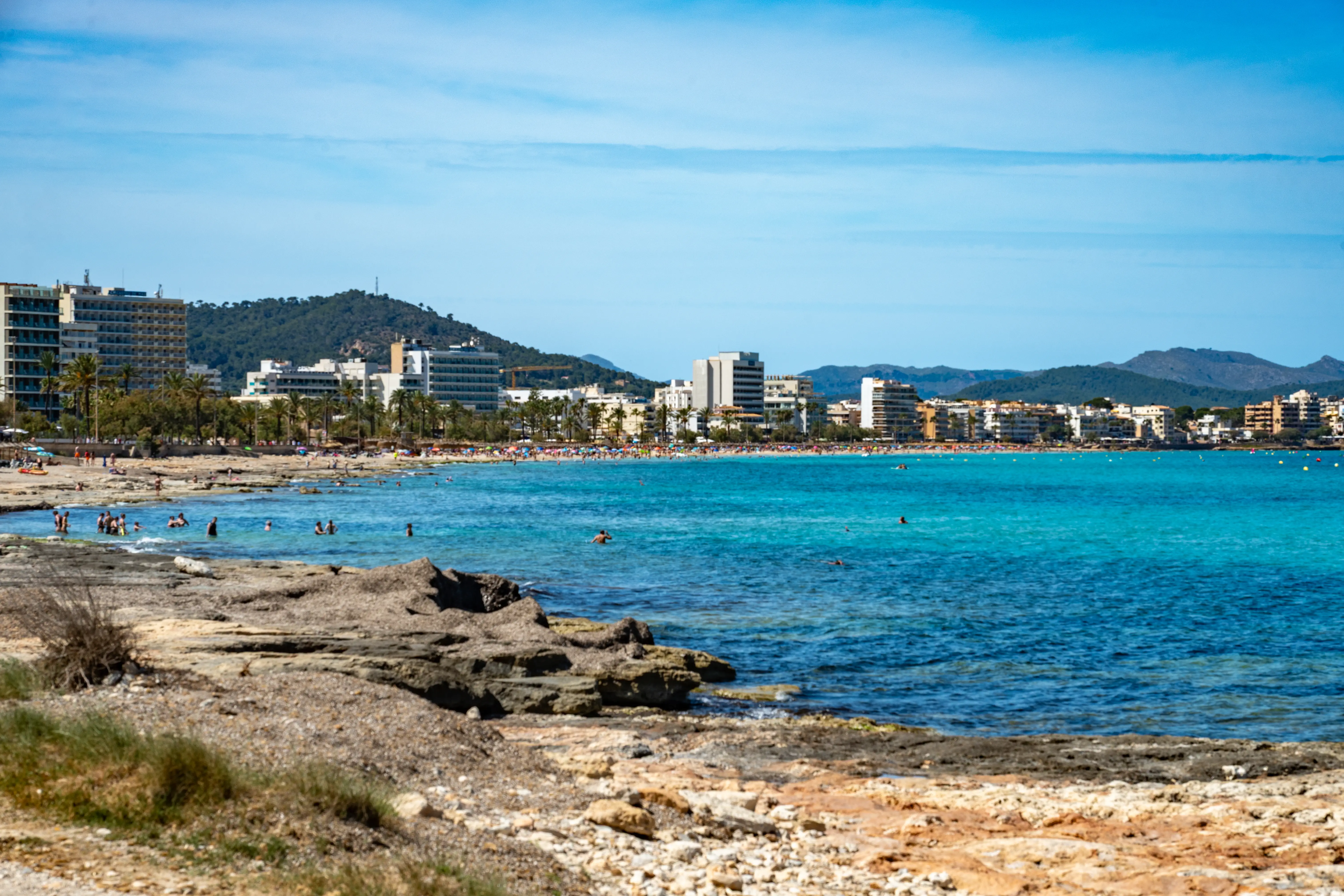 A panoramic view of a bustling Mediterranean beach with shallow, turquoise water lapping against a rocky and sandy shore. Numerous tourists are visible swimming and relaxing on the beach, with a line of hotels and apartment buildings forming the backdrop, all set against a backdrop of lush green hills and a clear blue sky.