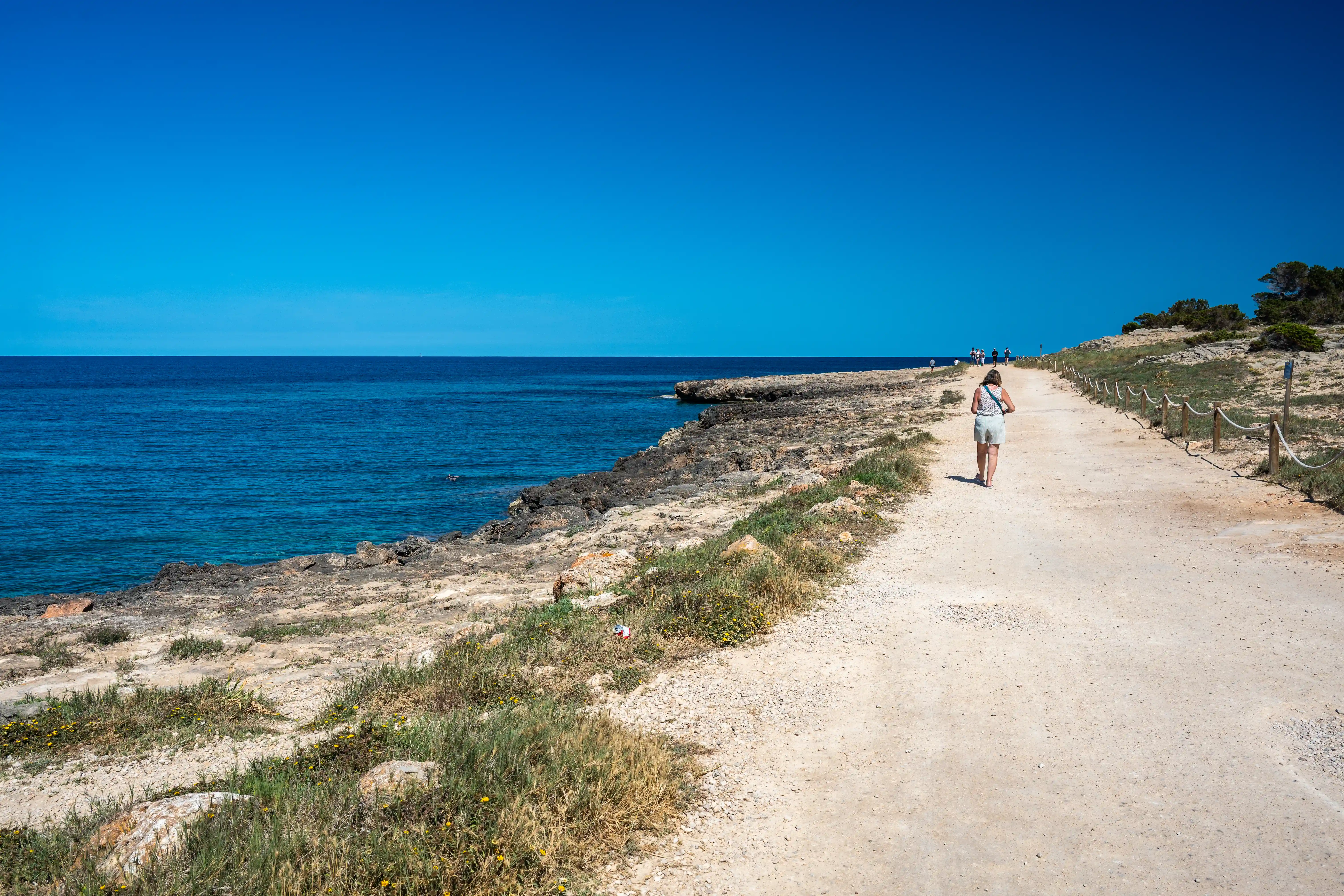 A woman in a light-coloured dress walks away from the camera on a sandy path bordering a rocky Mediterranean coastline. The sea is a vibrant blue and the sky is clear.