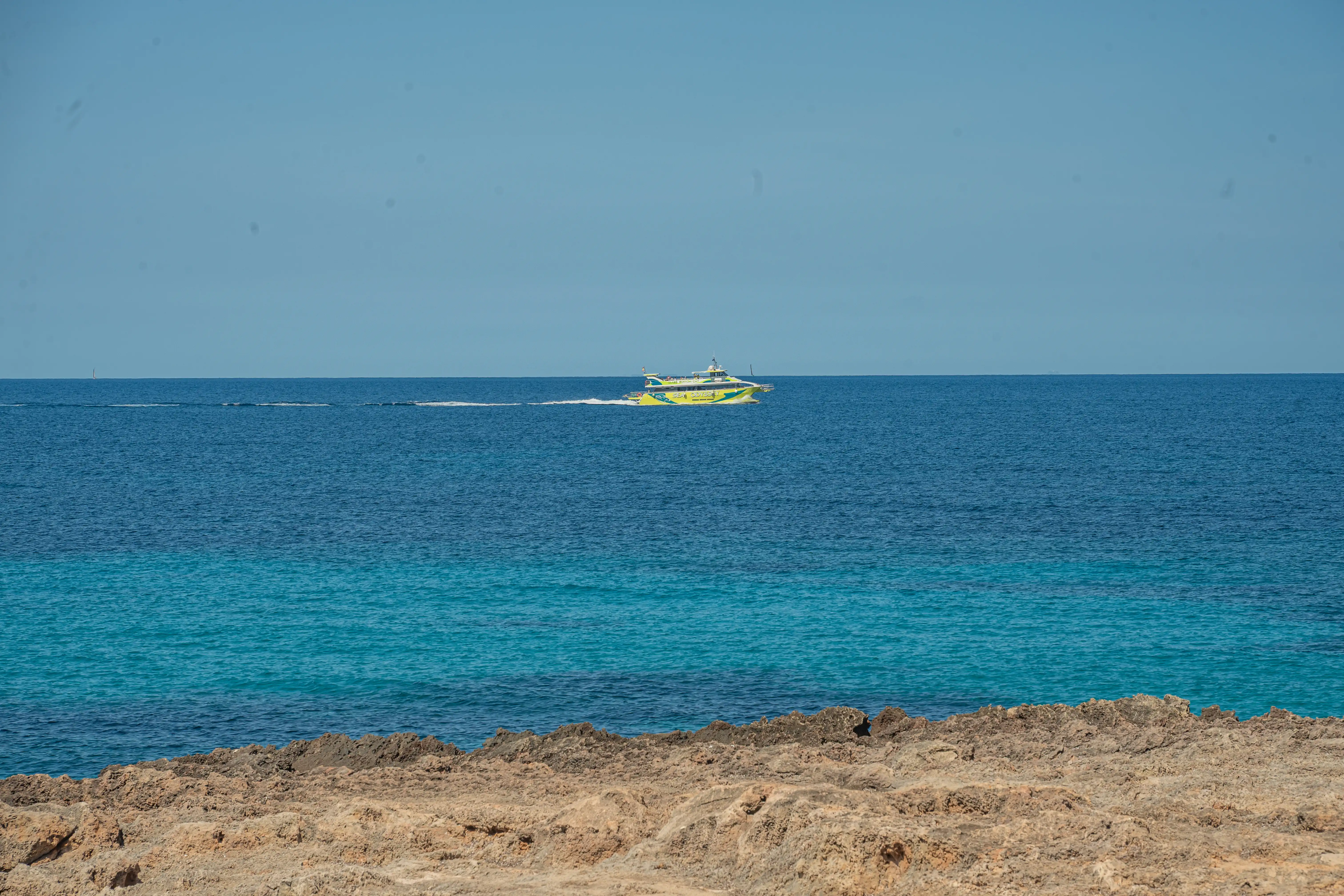 A bright yellow and green hydrofoil ferry travels on the ocean. The water is a mix of deep blue and turquoise, with a rocky shore in the foreground under a clear blue sky.