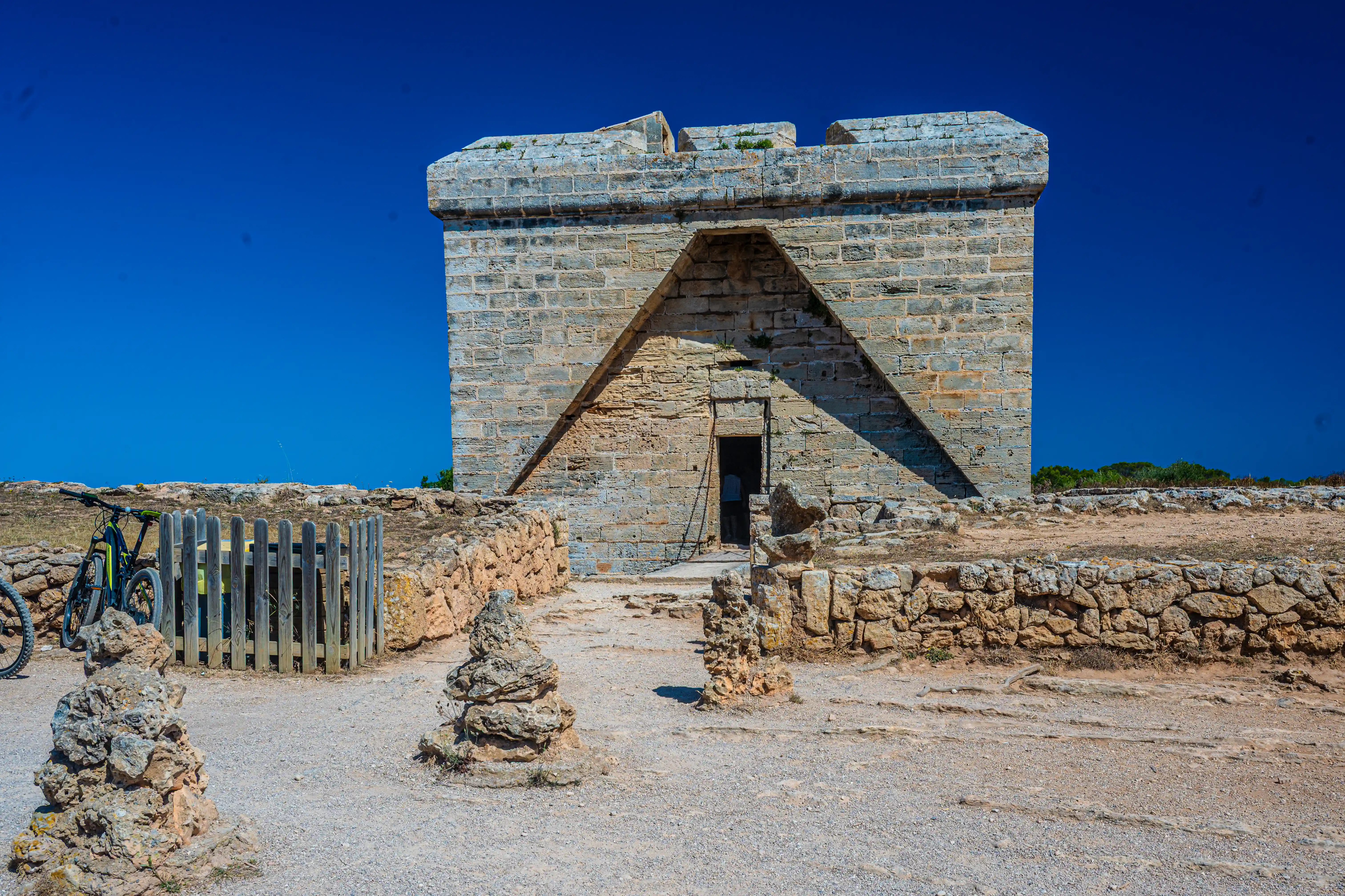 A historic stone watchtower with a triangular entrance, set against a bright blue sky. A bicycle is parked to the left, and stacked stones are visible in the foreground.