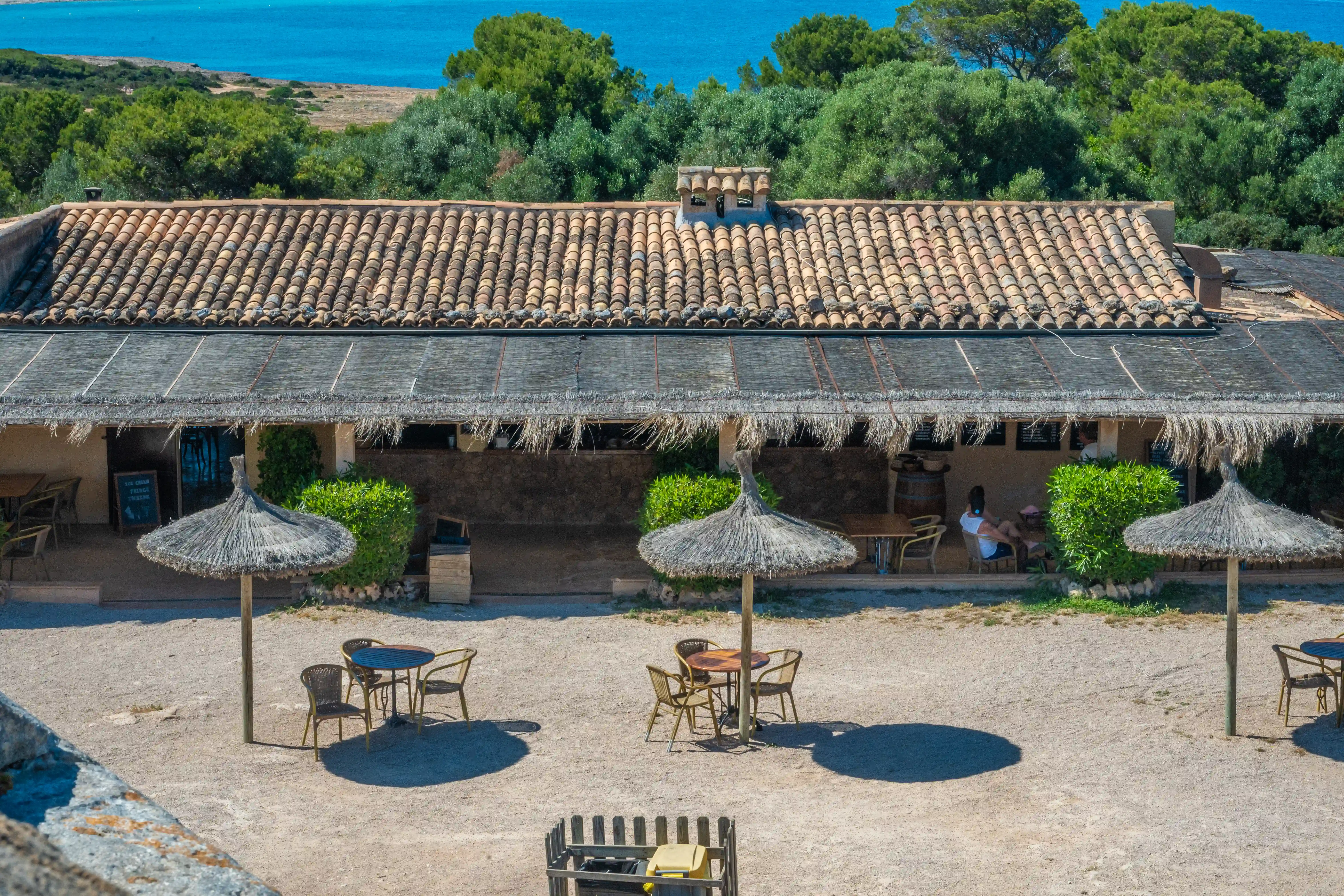 A seaside restaurant with a traditional tiled roof and a thatched outdoor seating area, featuring straw umbrellas and tables on a sandy terrace, with green foliage and the blue sea in the background.
