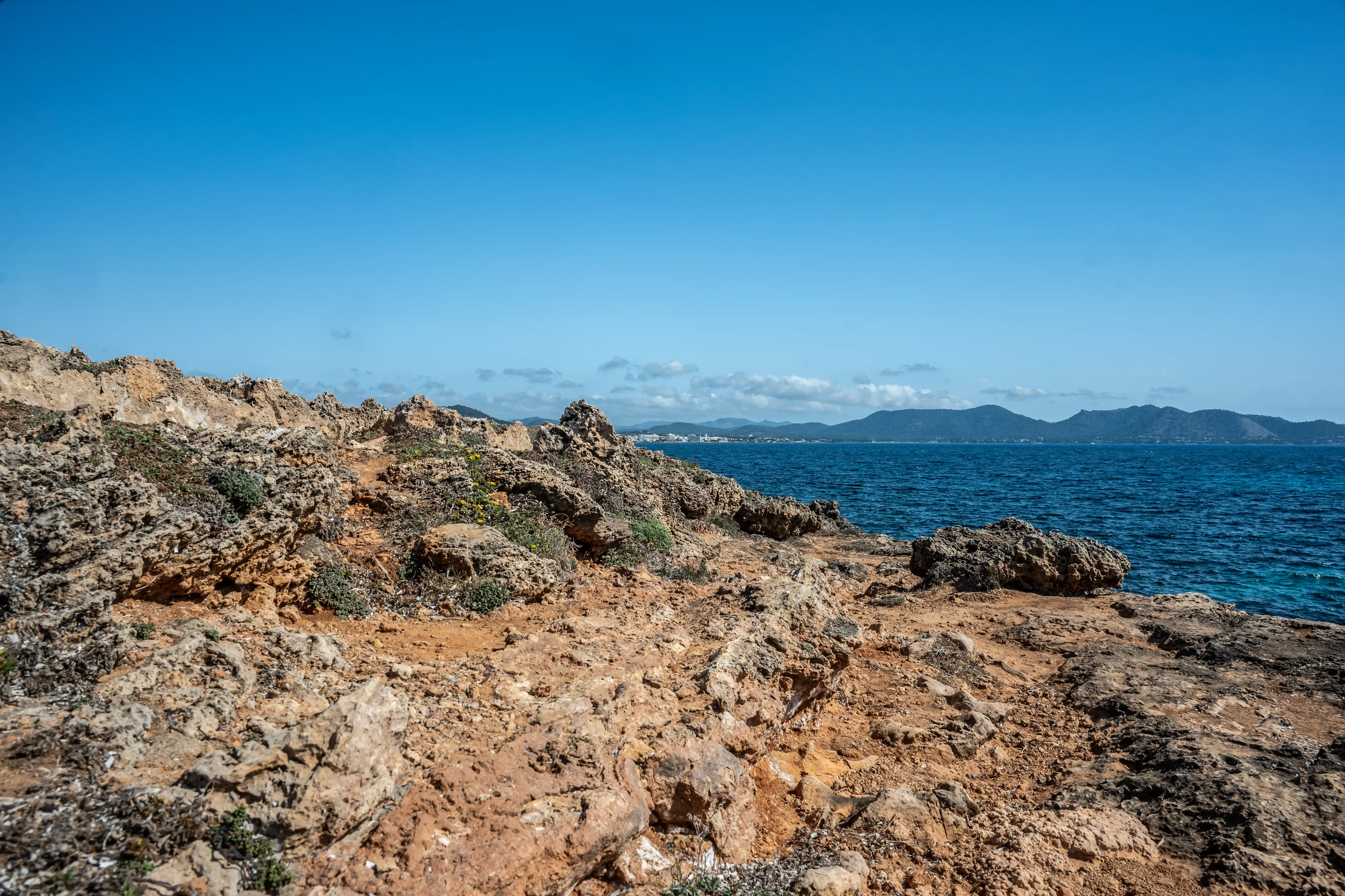 A sun-drenched, rocky shoreline meets the deep blue sea, with a hazy mountainous landscape in the distance under a clear blue sky.