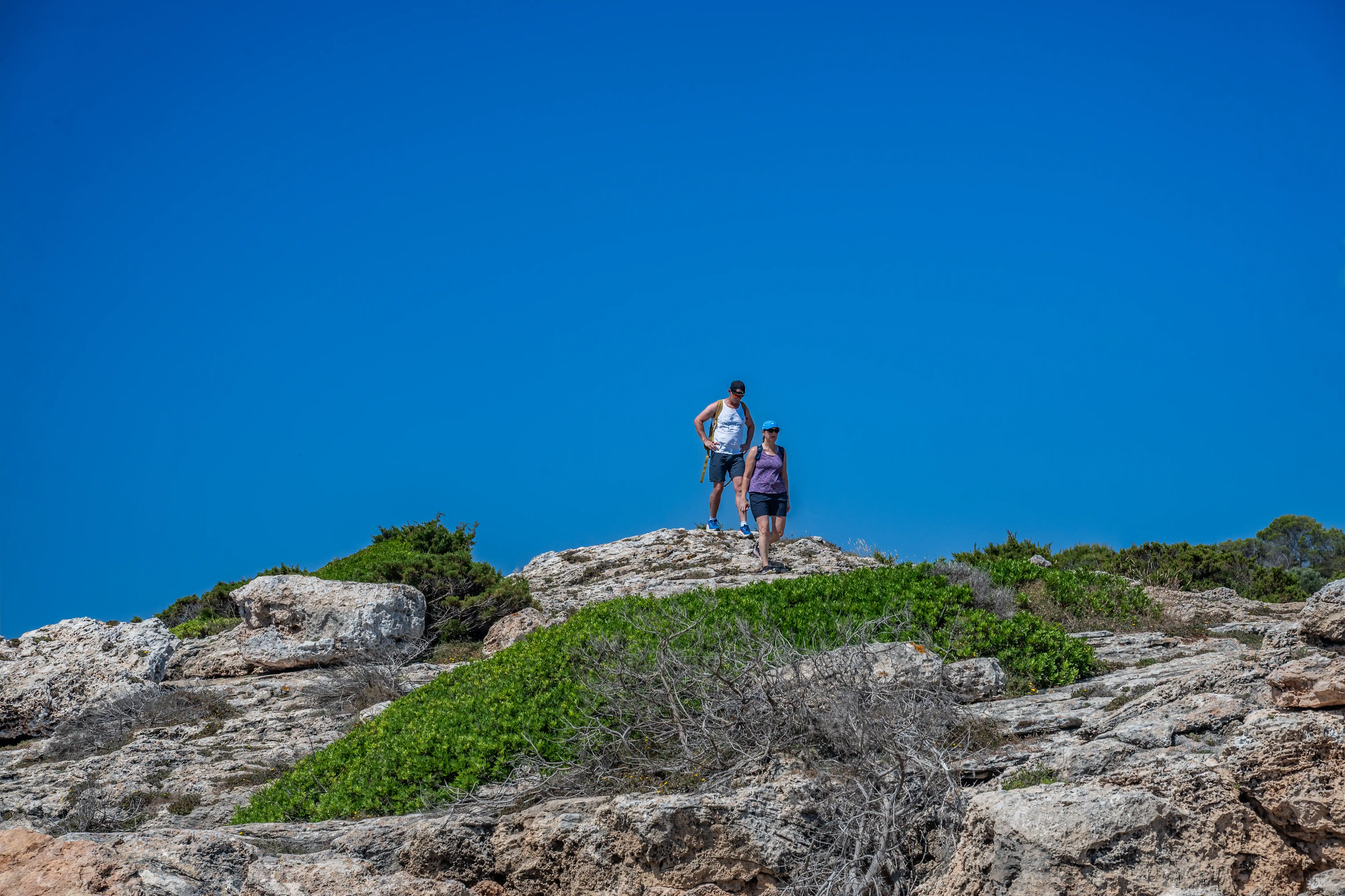 Two hikers, a man in a white tank top and shorts, and a woman in a purple top and dark shorts, stand on a rocky hill. The sky is a vibrant, clear blue.