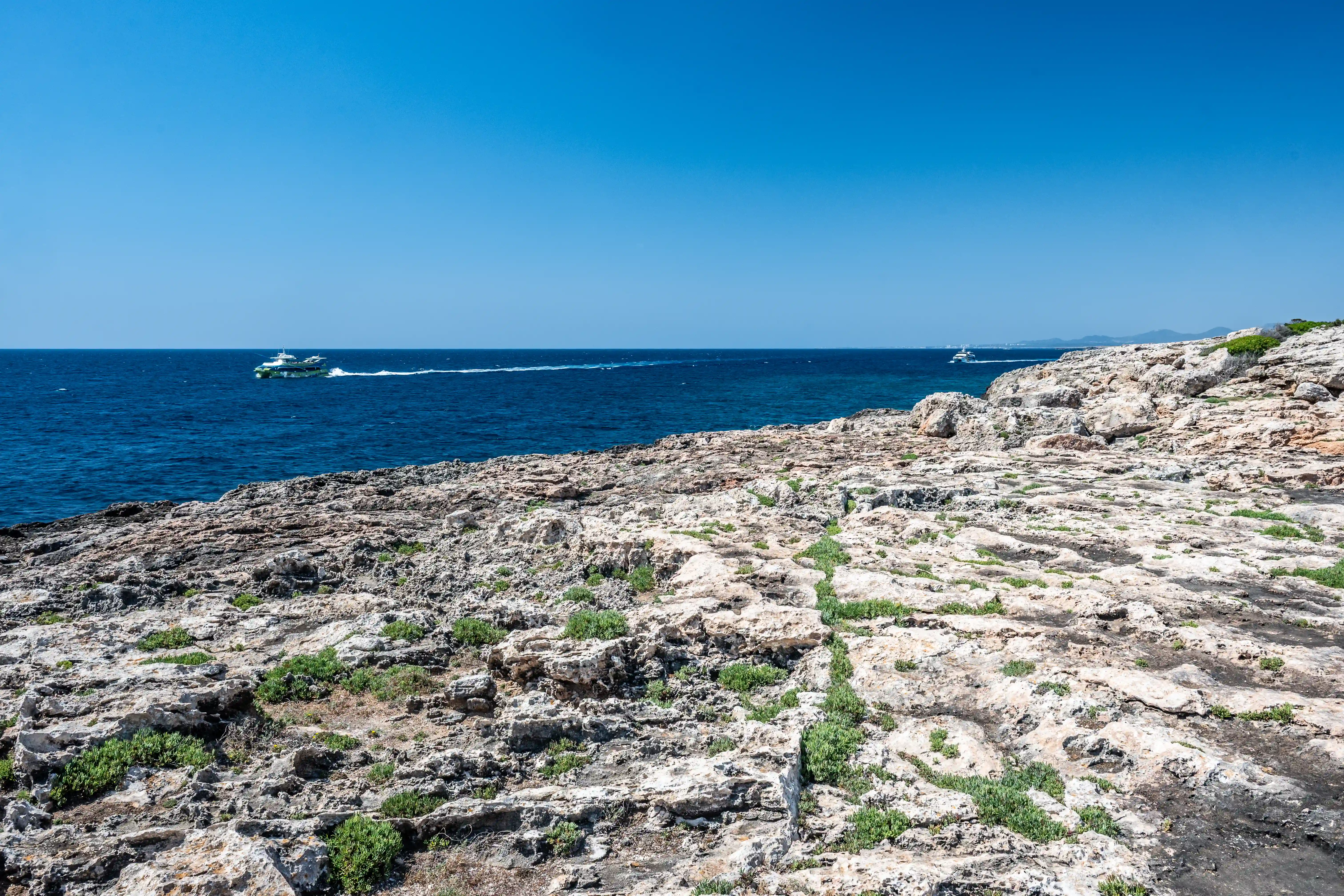 A rocky Mediterranean coastline with sparse green vegetation under a clear blue sky. Two white boats are sailing on the dark blue sea in the distance.
