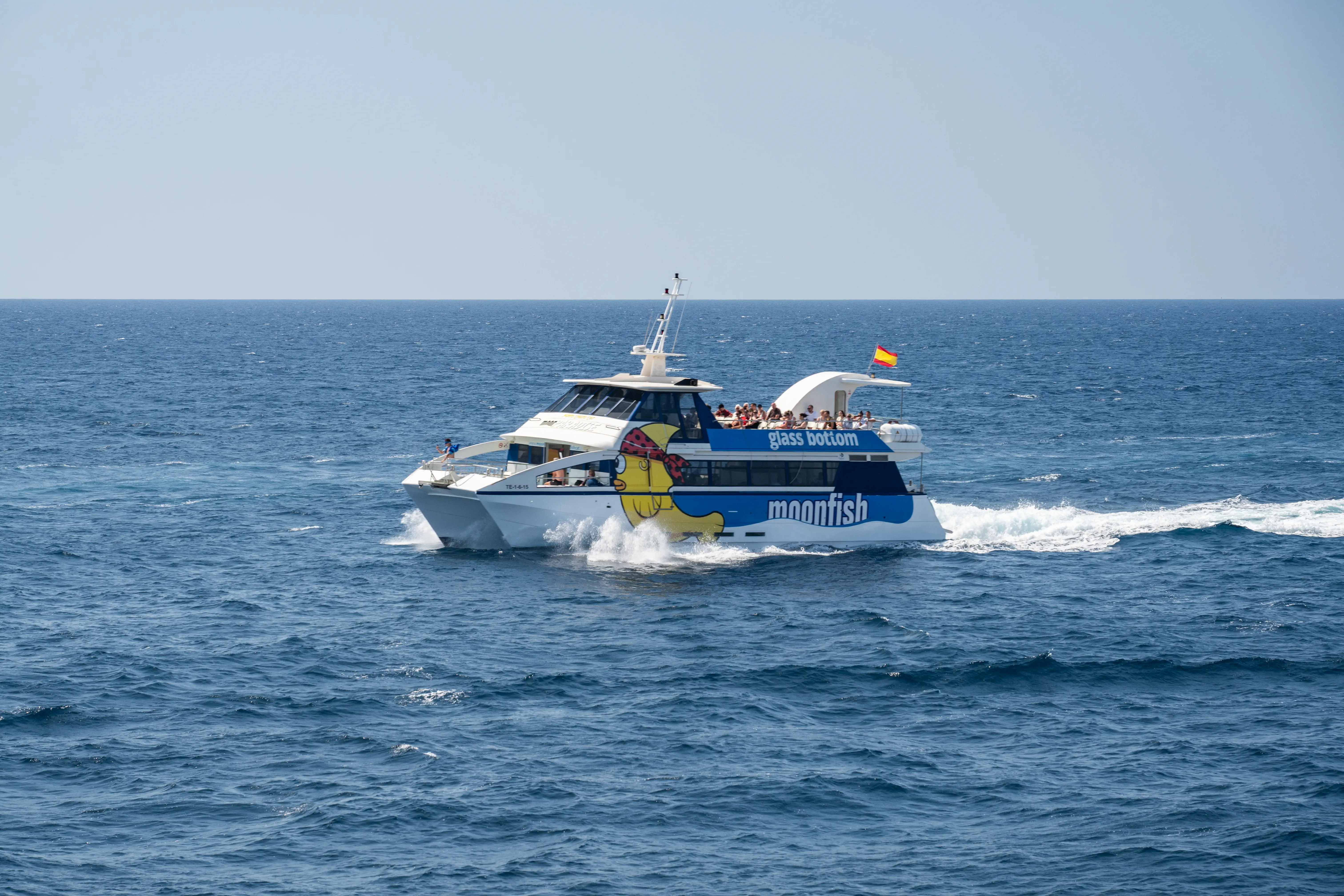A glass bottom boat named Moonfish with passengers on its upper deck sails through blue ocean water on a sunny day.