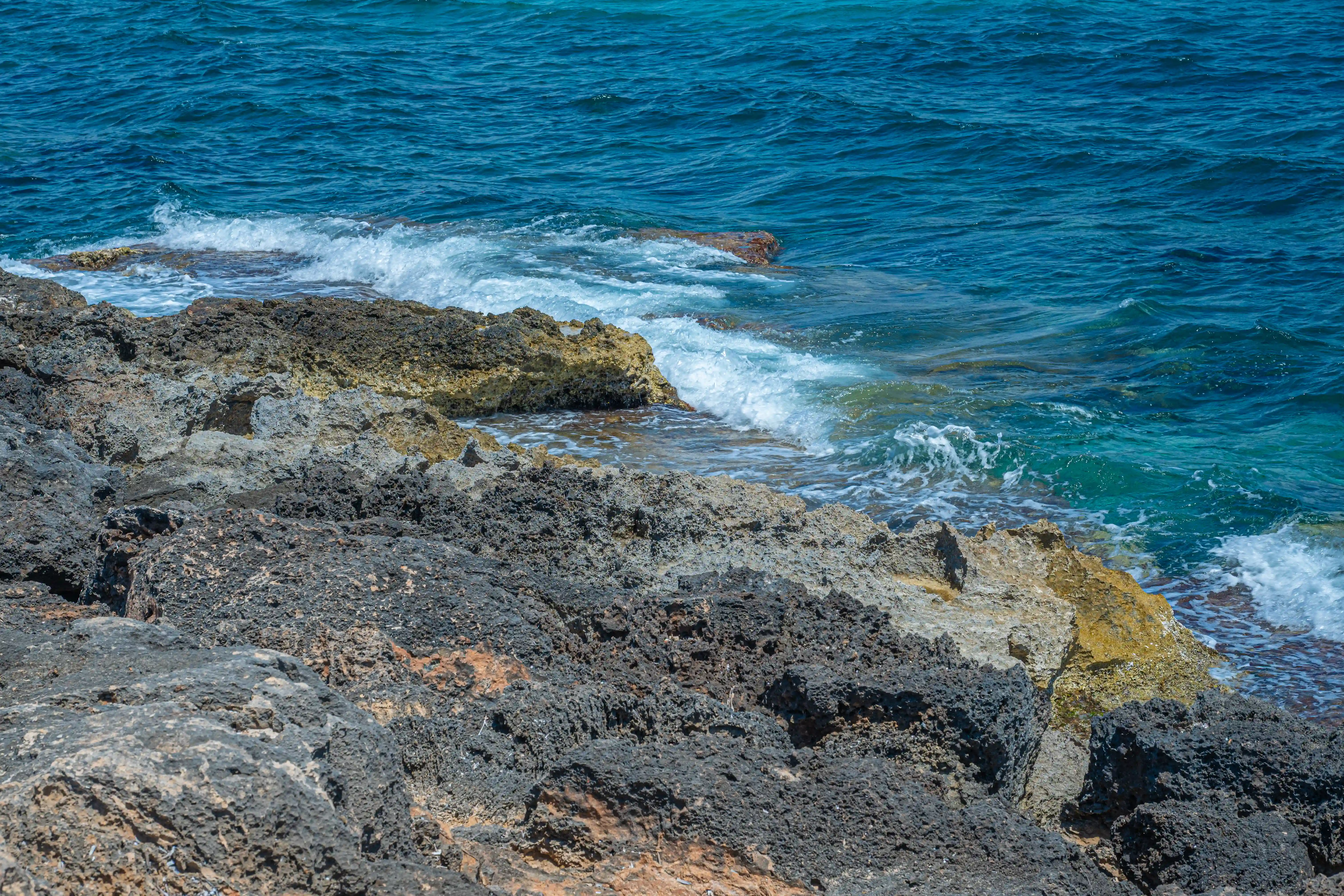 Close-up of dark, porous volcanic rocks on a shoreline, with bright turquoise sea water and white-capped waves breaking against the rocks.