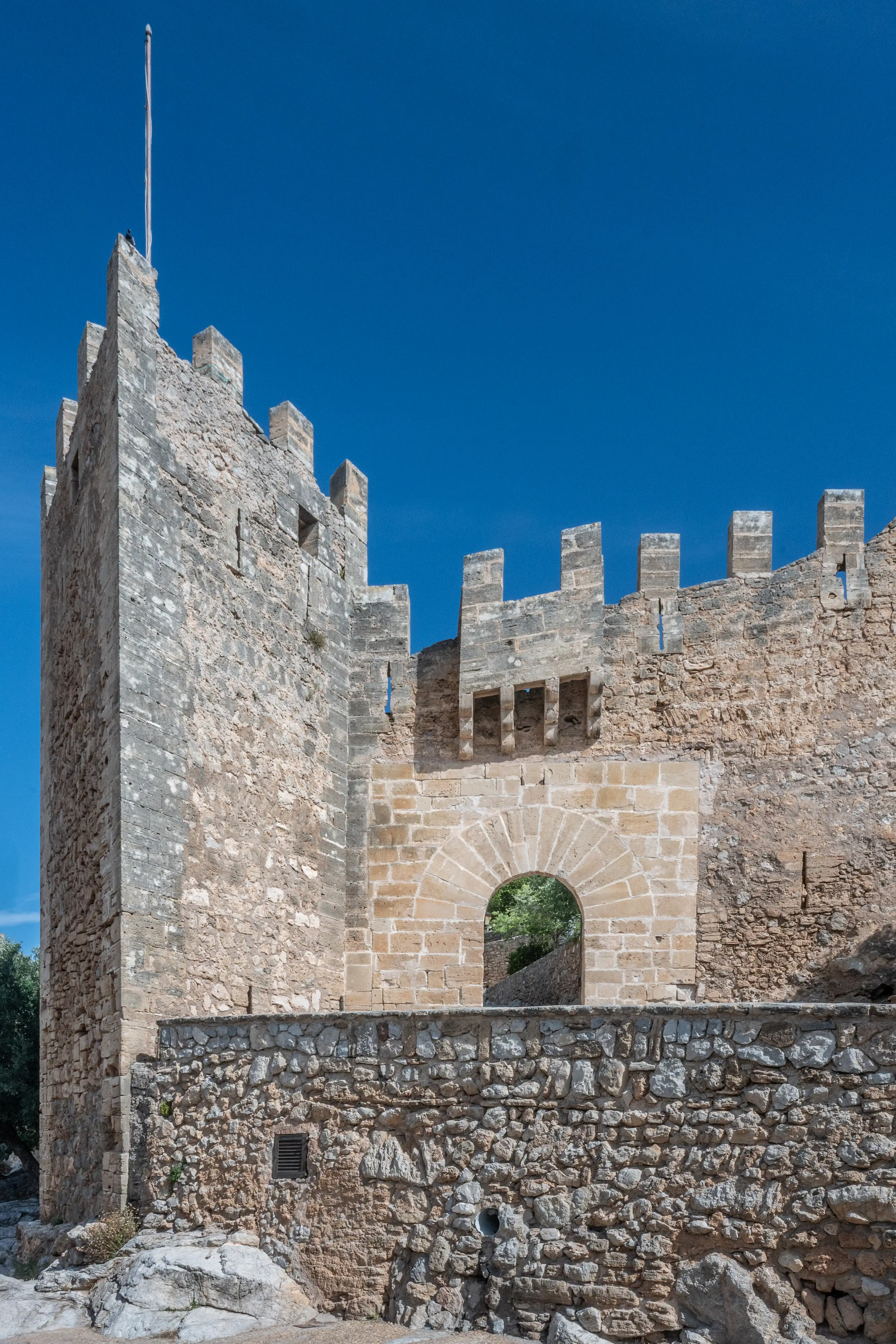 Close-up of a medieval stone castle tower and wall with battlements and an arched entrance against a vibrant blue sky.