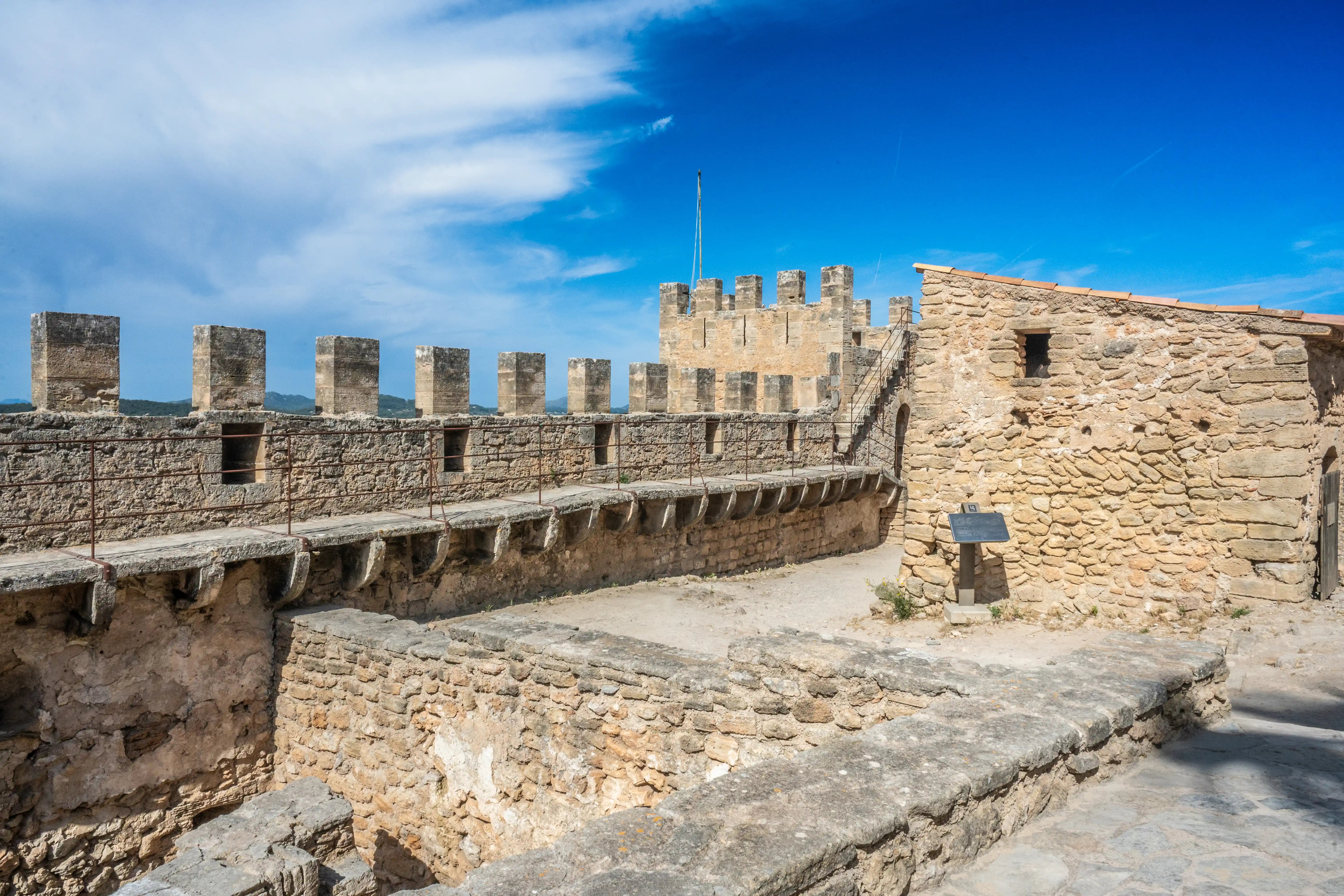 Stone battlements of an old castle with crenellations and narrow windows, a walkway with metal railings, a stone building, and a bright blue sky with white clouds.