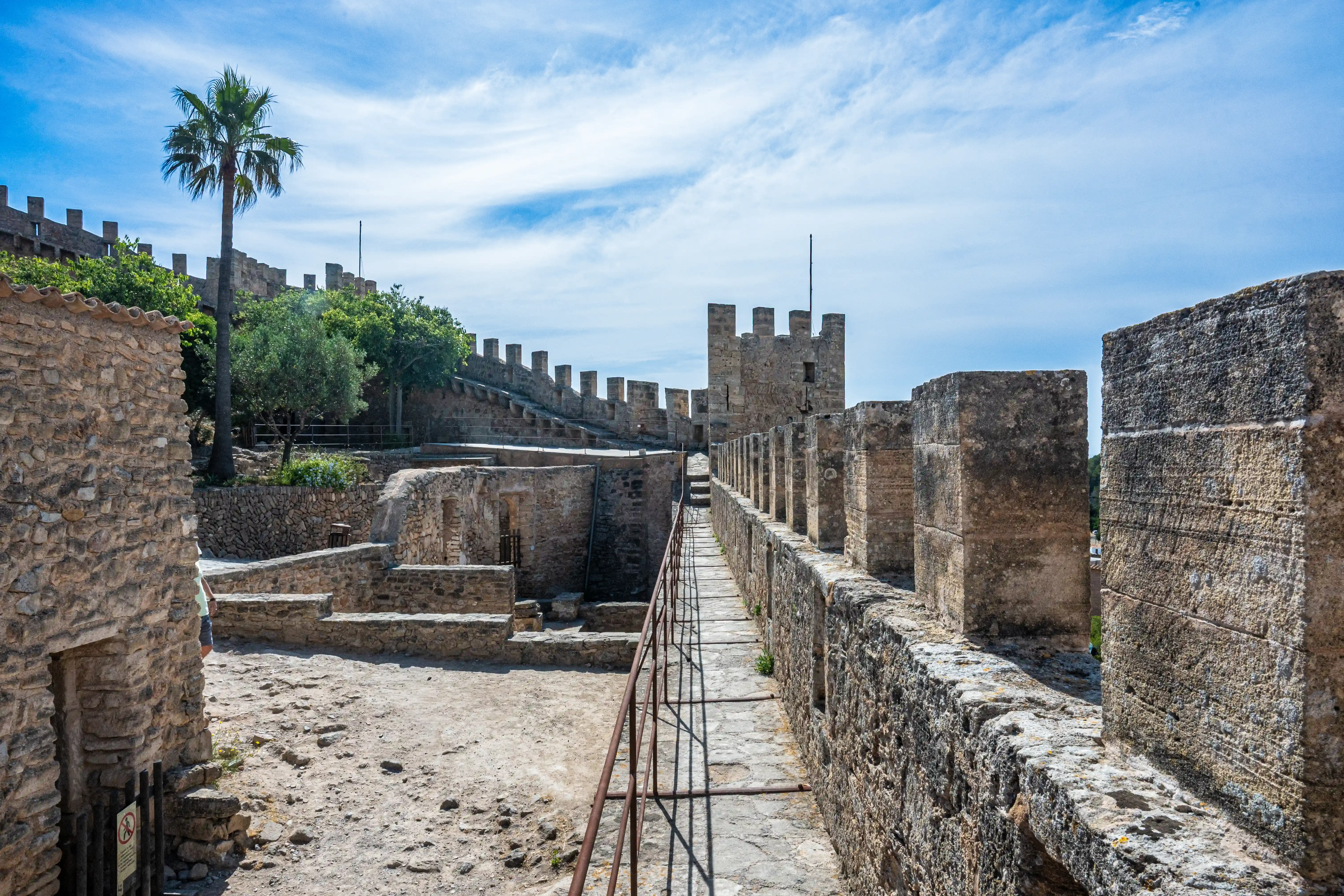 A scenic view of a historic castle's stone fortifications under a bright blue sky, featuring a prominent palm tree and the textured detail of the ancient walls.